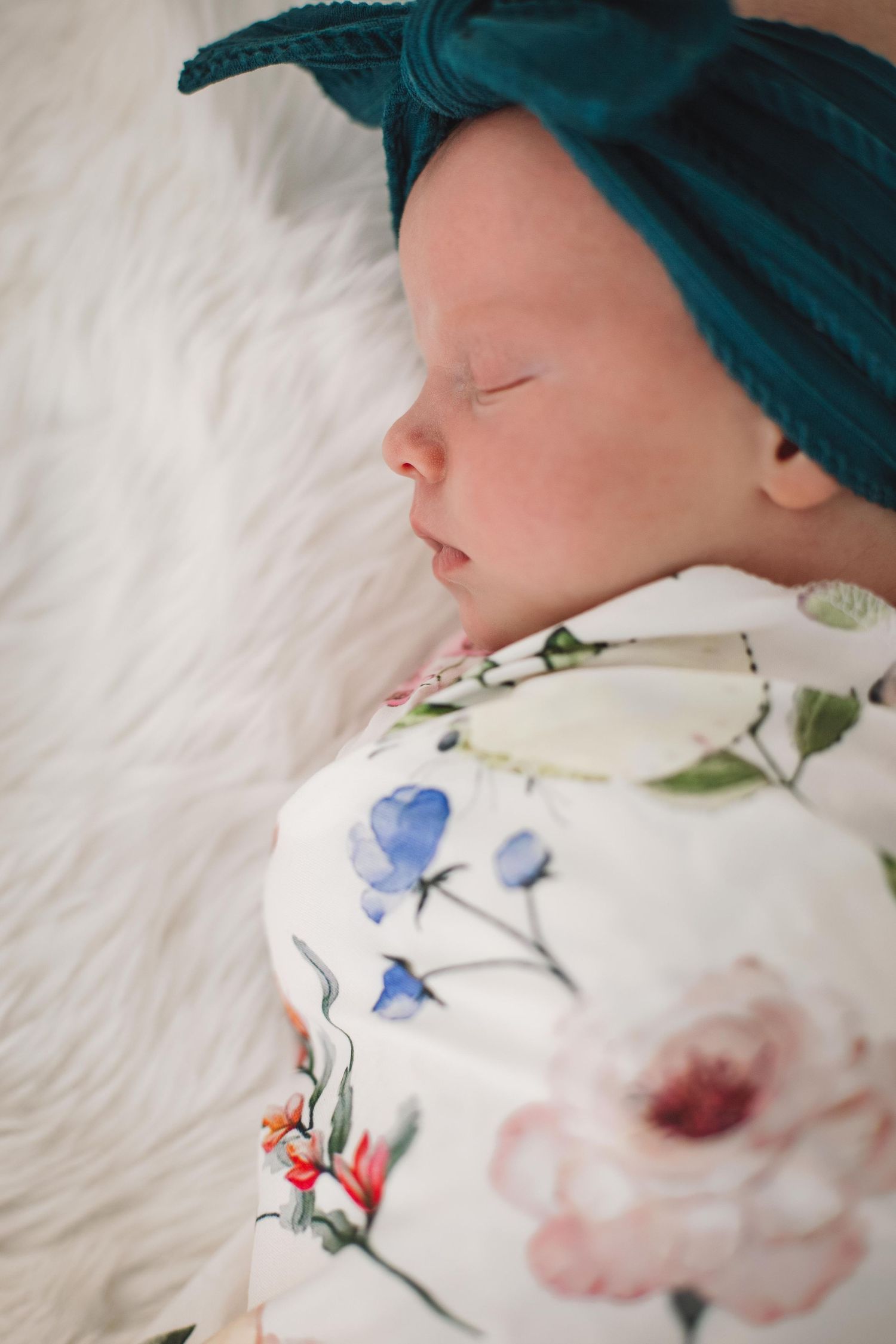 Sleeping infant in teal headband and floral outfit on white fur backdrop.