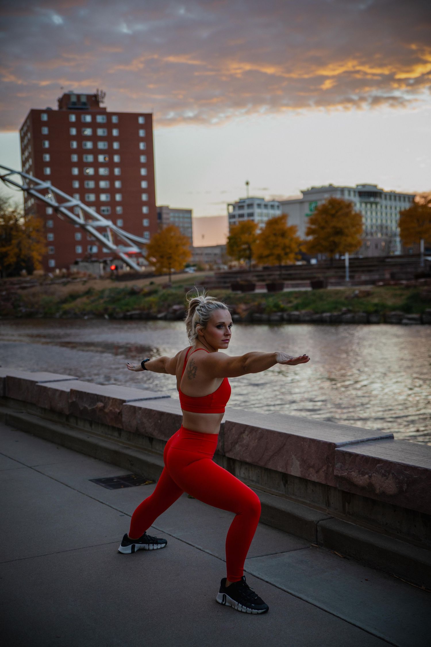 A person in bright red athletic wear performs yoga poses along a waterfront at sunset with brick buildings in background.