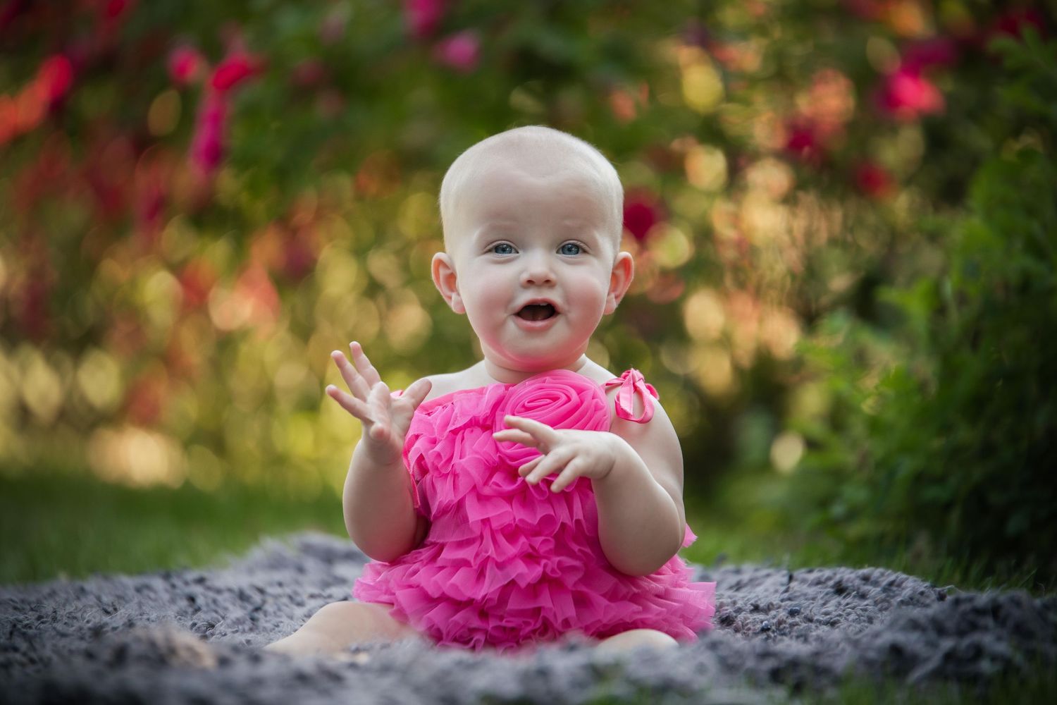 Baby wearing pink ruffled outfit shows excitement while sitting outdoors.