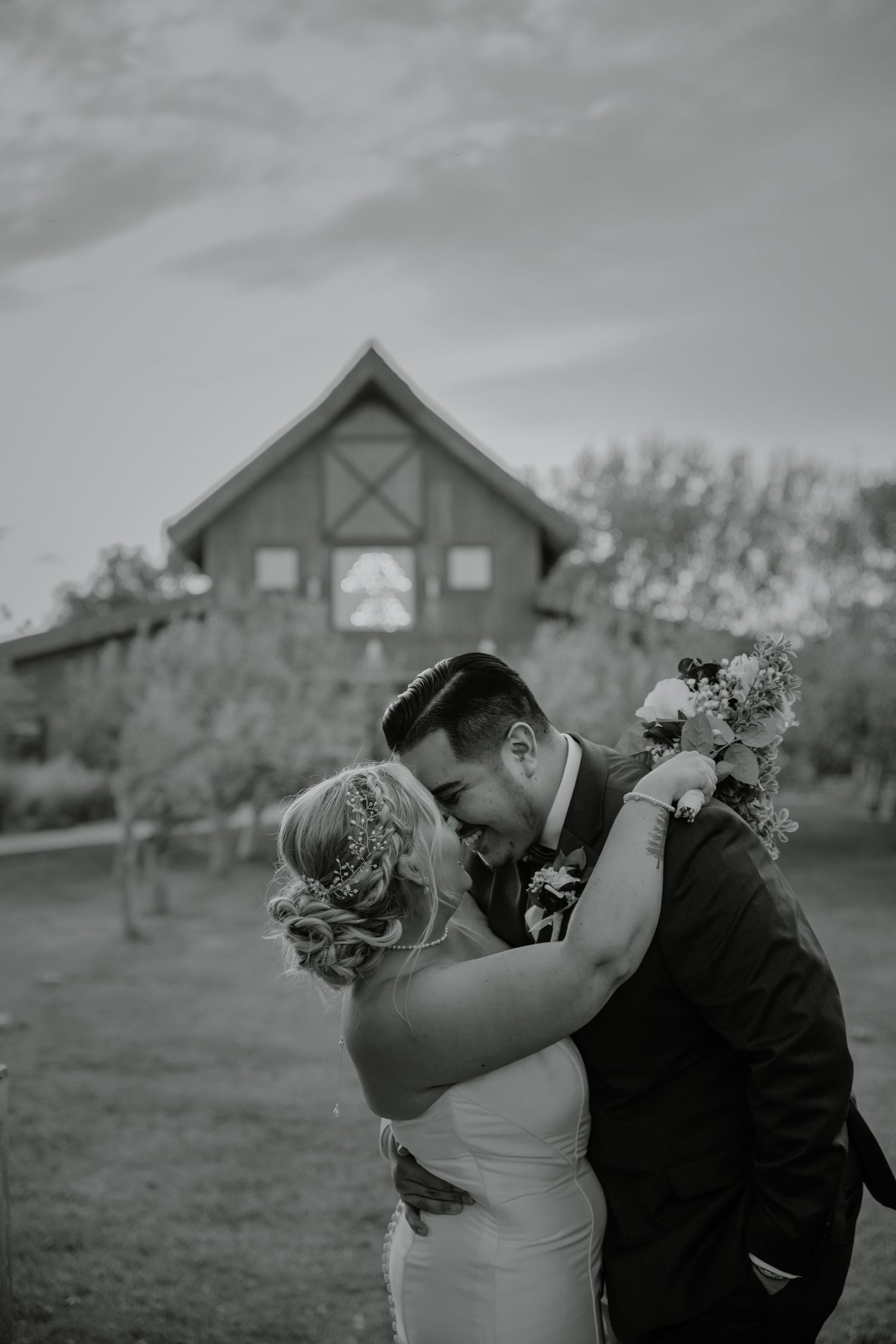 Black and white portrait of a romantic wedding moment with barn in background.