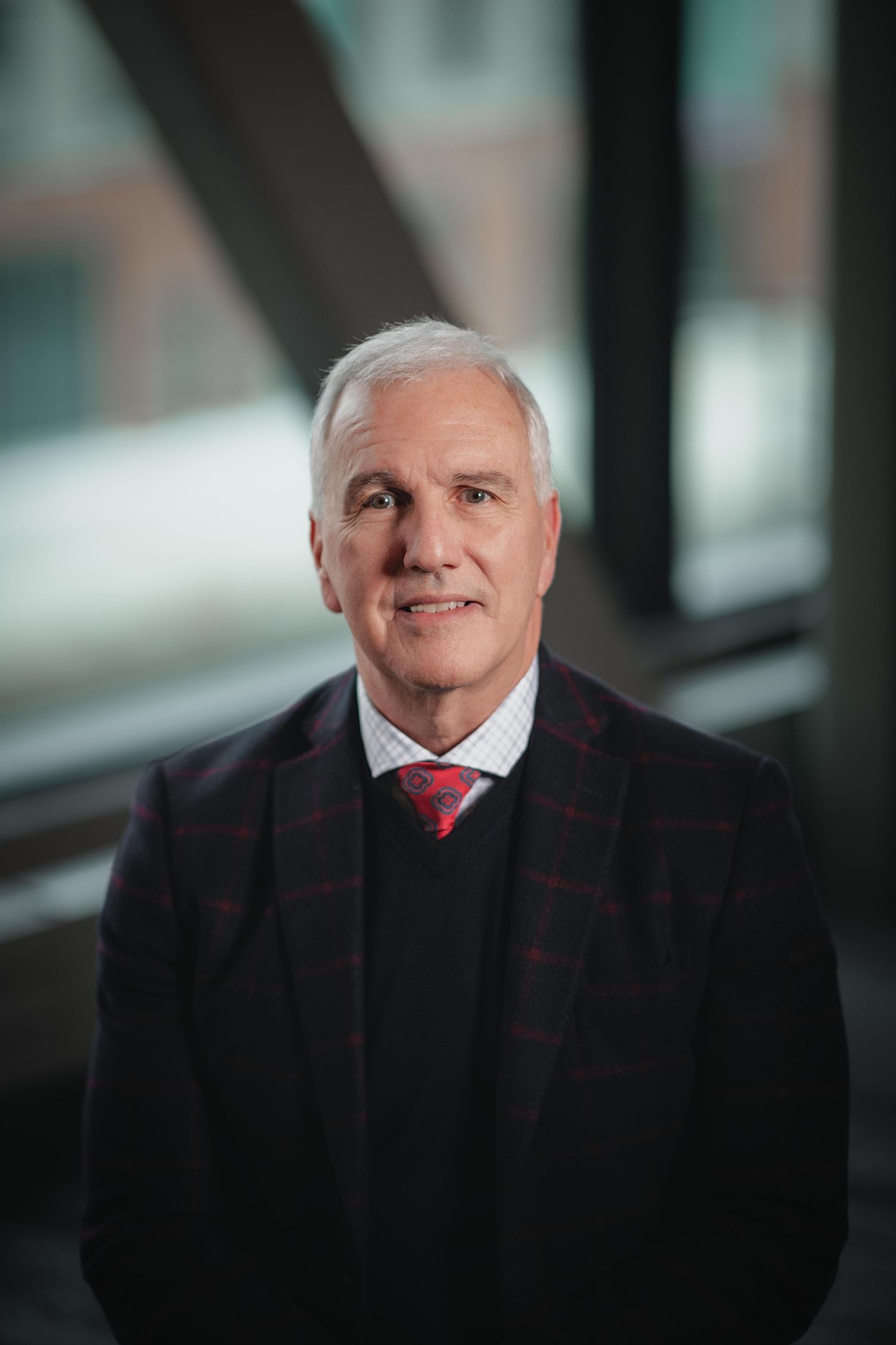 Professional portrait of a senior executive in a dark suit and red tie against a soft-focus background.