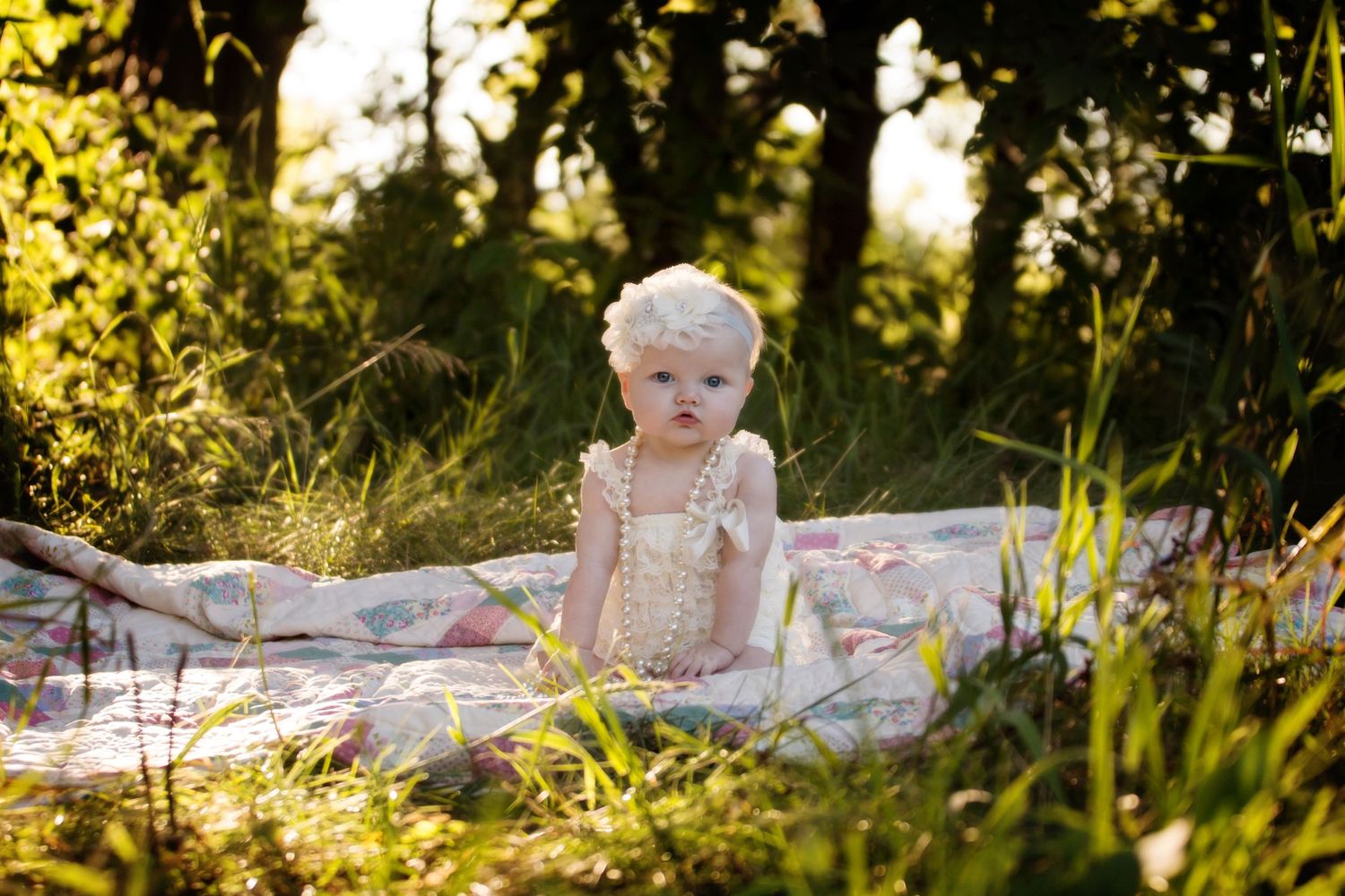 Baby in white dress sitting on blanket in sun-dappled summer grass.