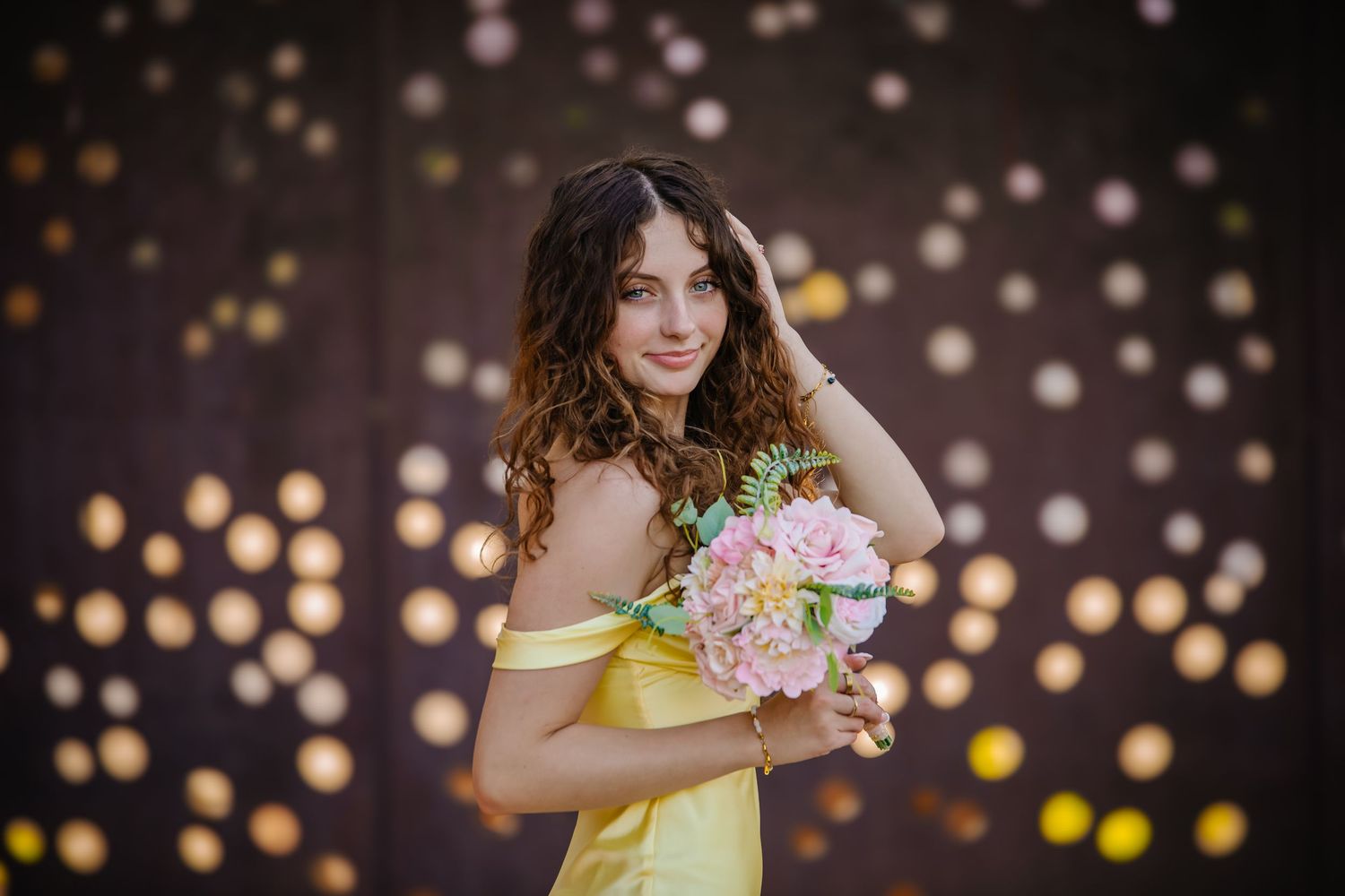 A woman in a yellow dress poses with pink flowers against a dark background with bokeh lights.