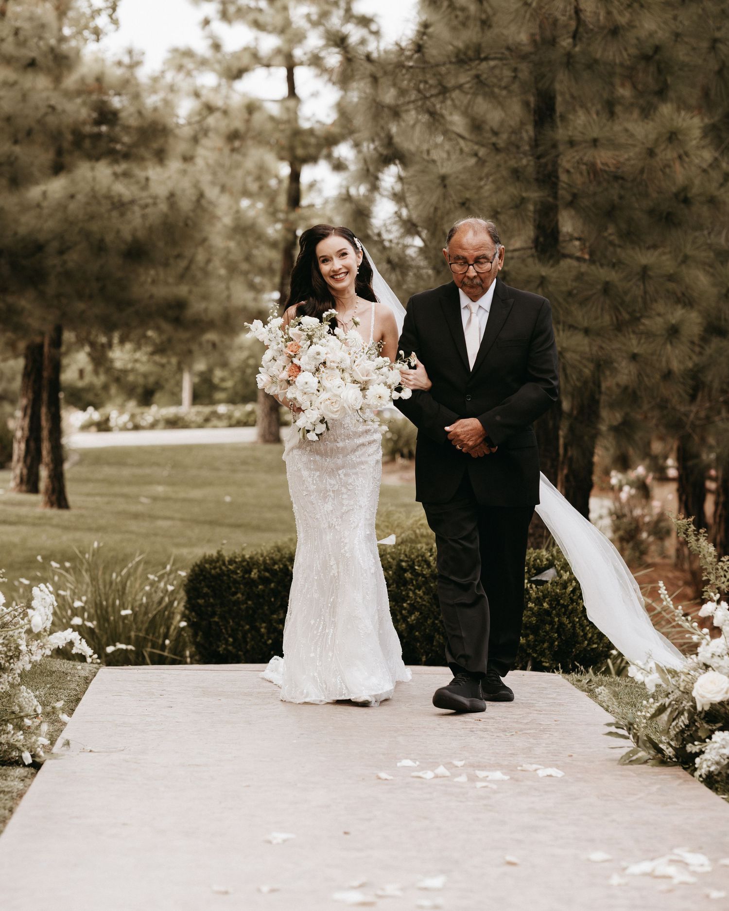 A bride in a fitted lace gown walks down a petal-lined path with an escort in a dark suit at an outdoor wedding ceremony.