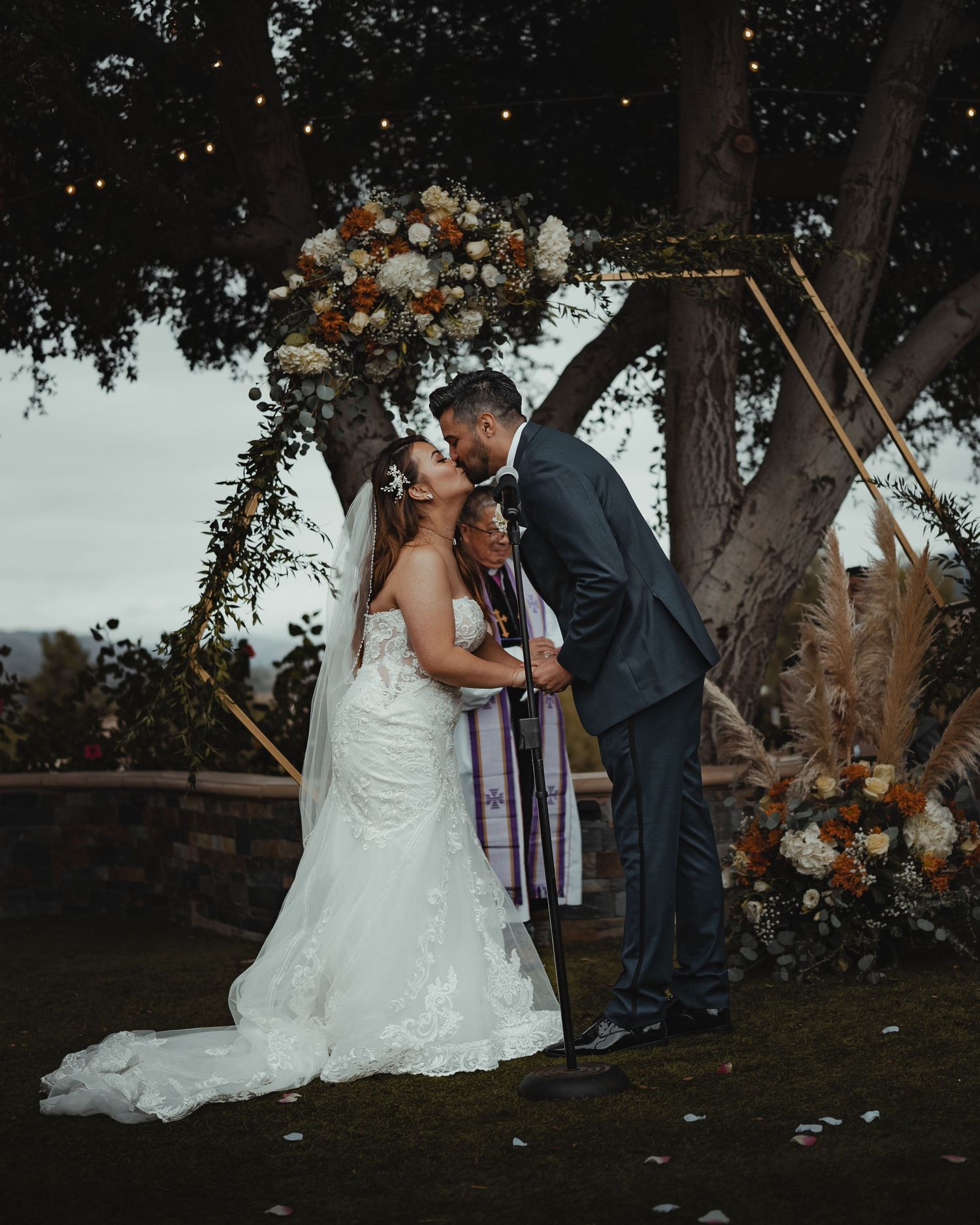 A couple shares an intimate moment during their outdoor wedding ceremony under trees at dusk.