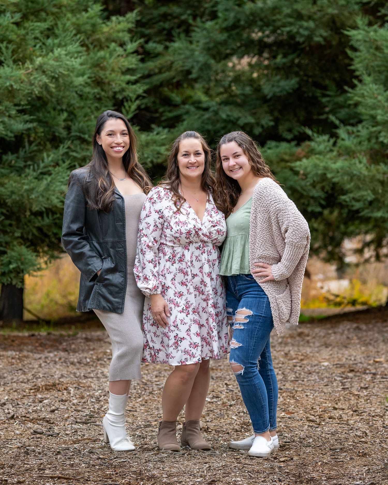 Three friends pose together outdoors on a fall day surrounded by green trees.