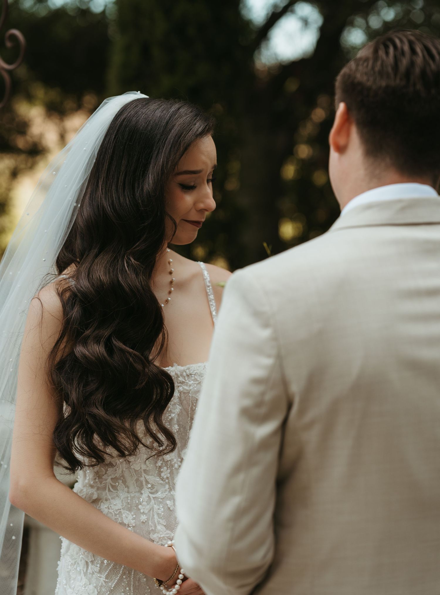 Bride with flowing dark wavy hair and embellished white gown stands with groom in beige suit during outdoor wedding ceremony.