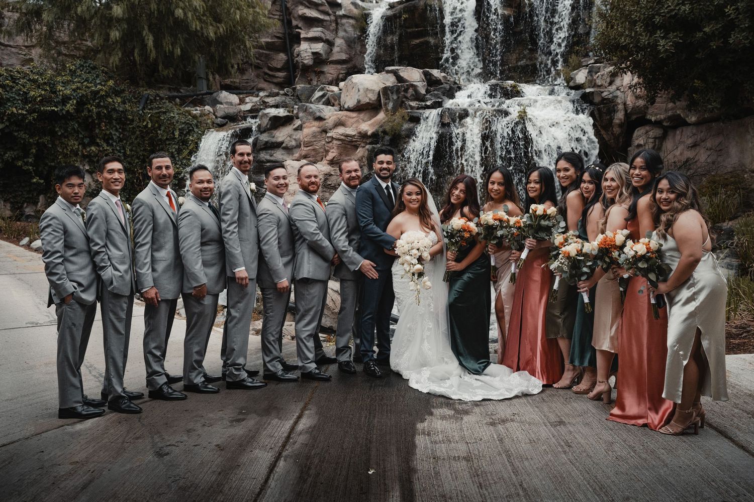Wedding party lined up by waterfall with bridesmaids in rust dresses and groomsmen in gray suits.