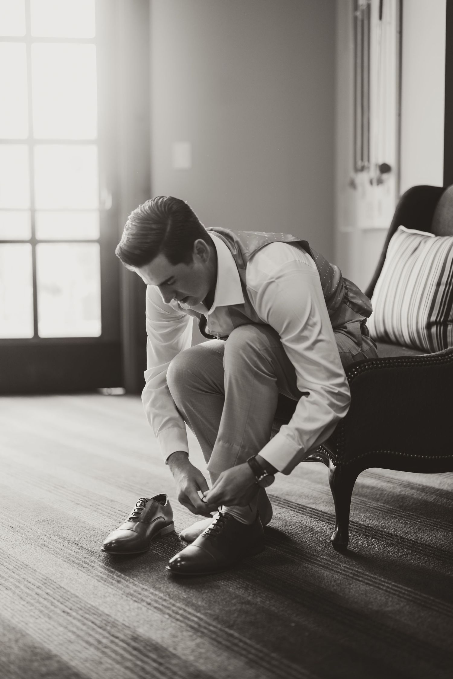 Black and white photo of person in formal attire tying dress shoes while seated on vintage chair by window.