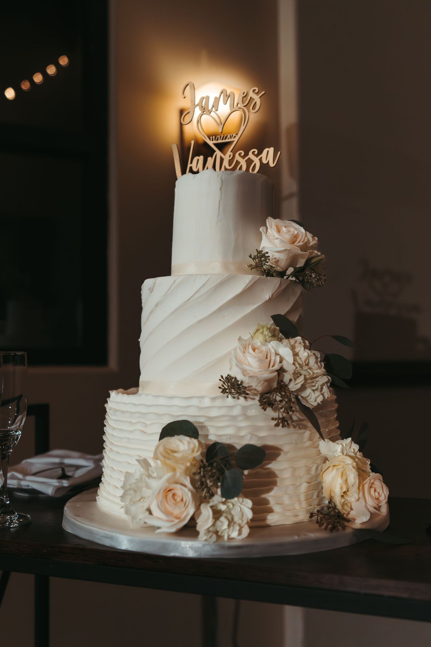 Elegant white wedding cake decorated with delicate roses and draped fondant layers illuminated by warm lighting.