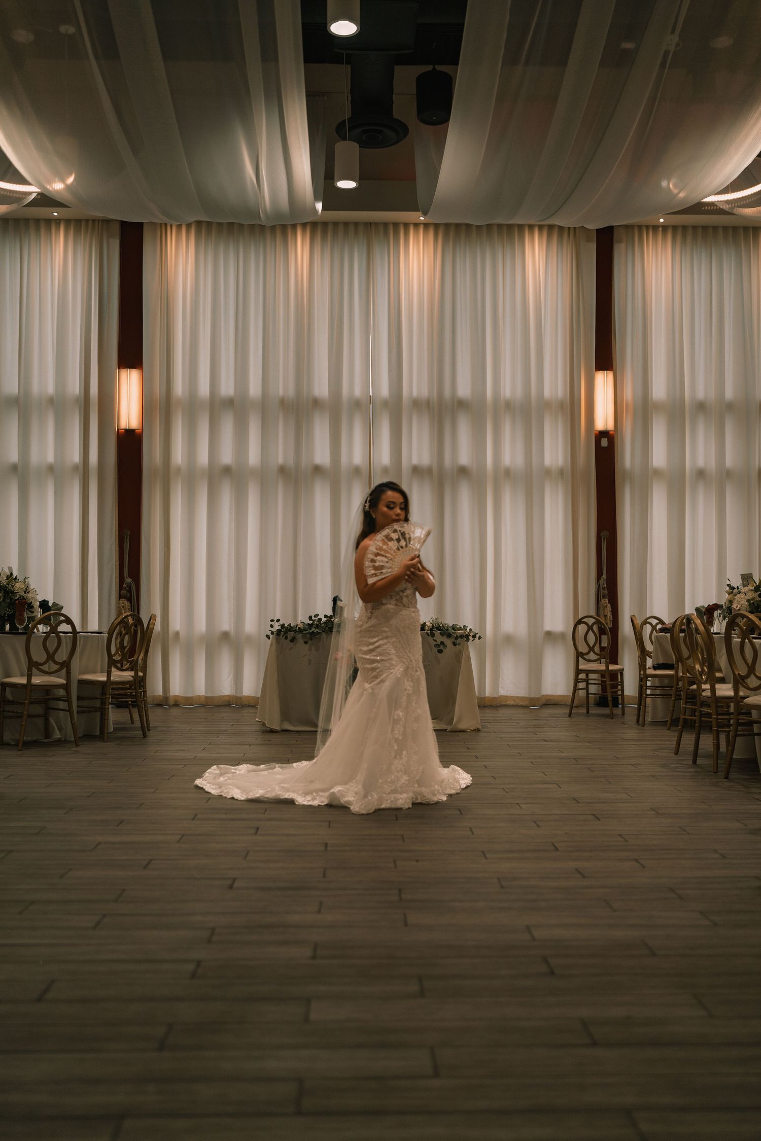 Spacious event venue with sheer curtains, wooden floors, and ambient lighting.