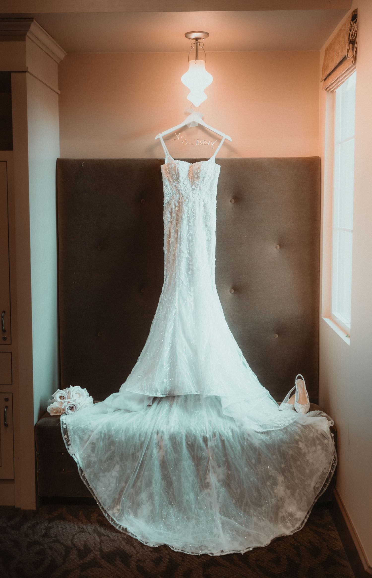 Wedding dress hanging in window light creating ethereal silhouette against neutral wall.