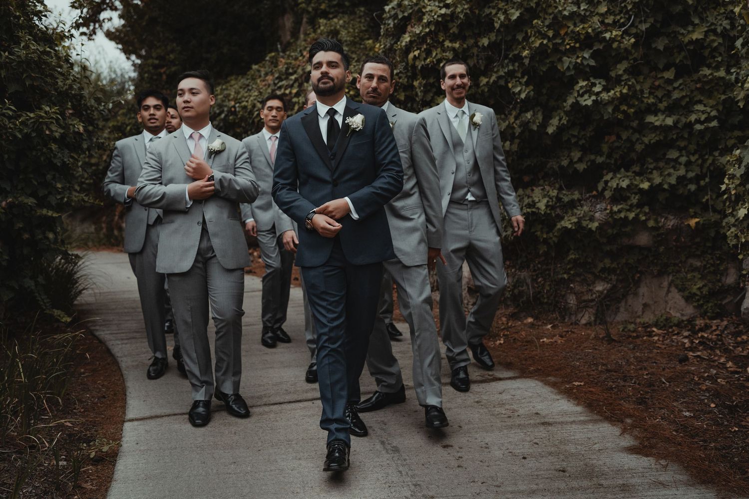 Wedding party in formal attire walking along landscaped garden path.