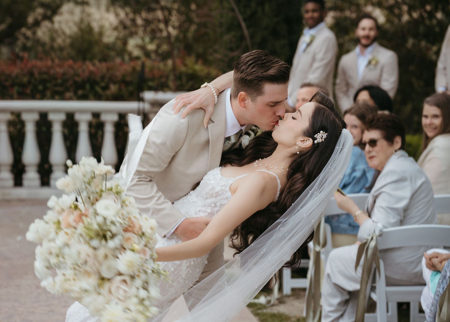 A couple shares a romantic kiss during their outdoor wedding ceremony with guests watching in the background.