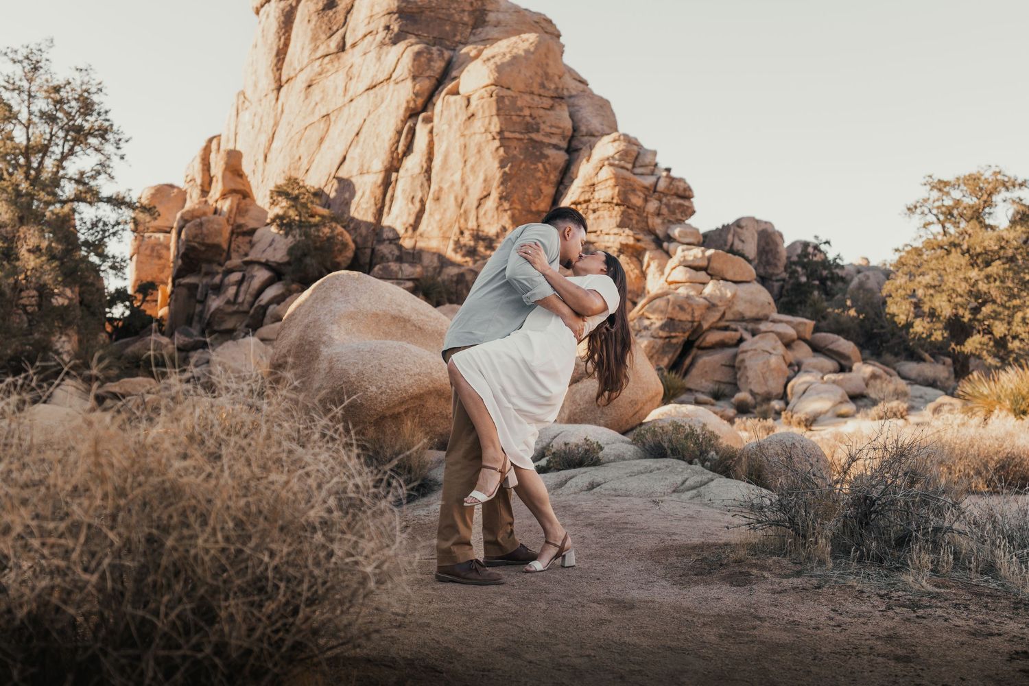 A couple poses for photos in a desert landscape with large rock formations at sunset.