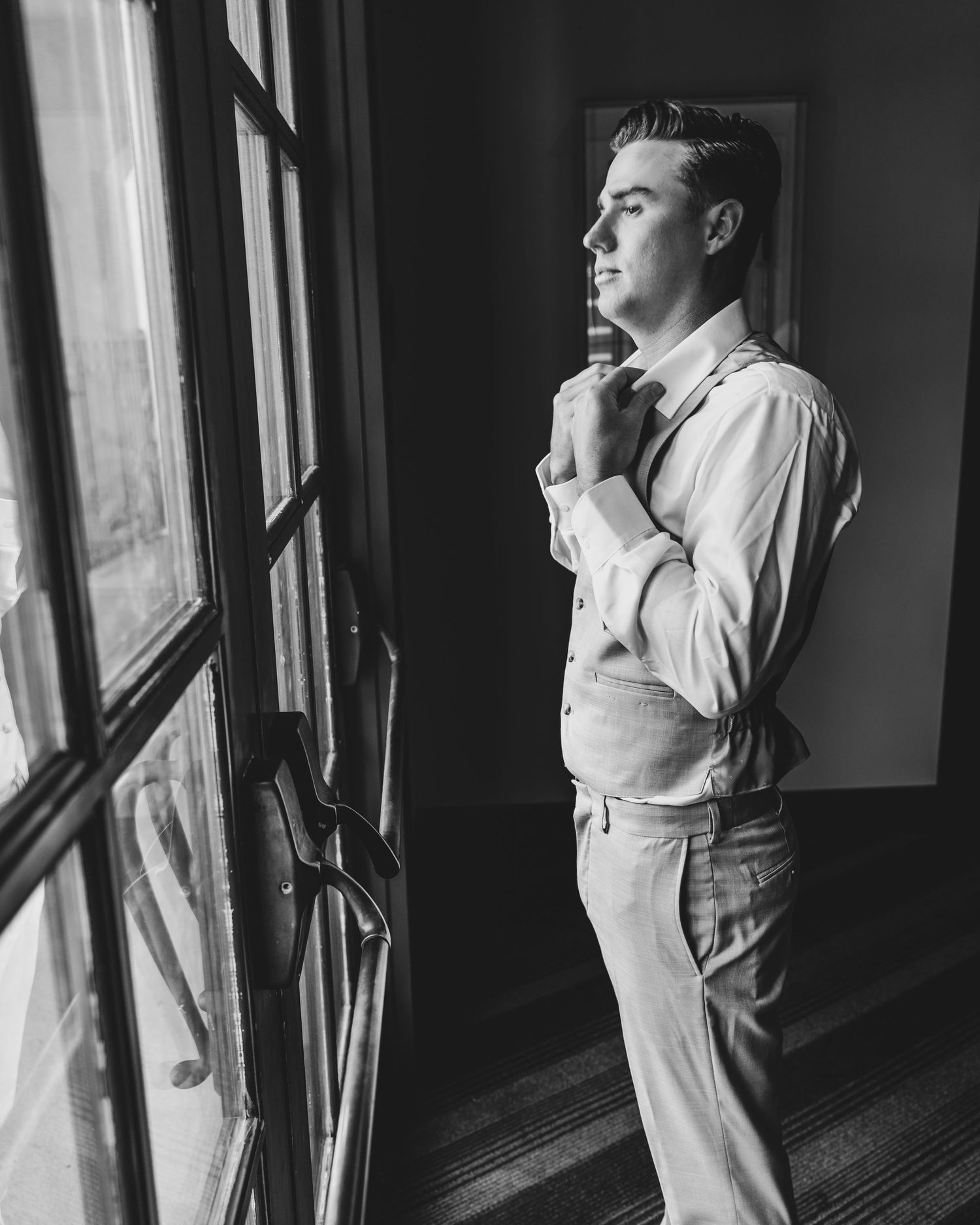 Classic black and white photo of a groom getting ready by a window.