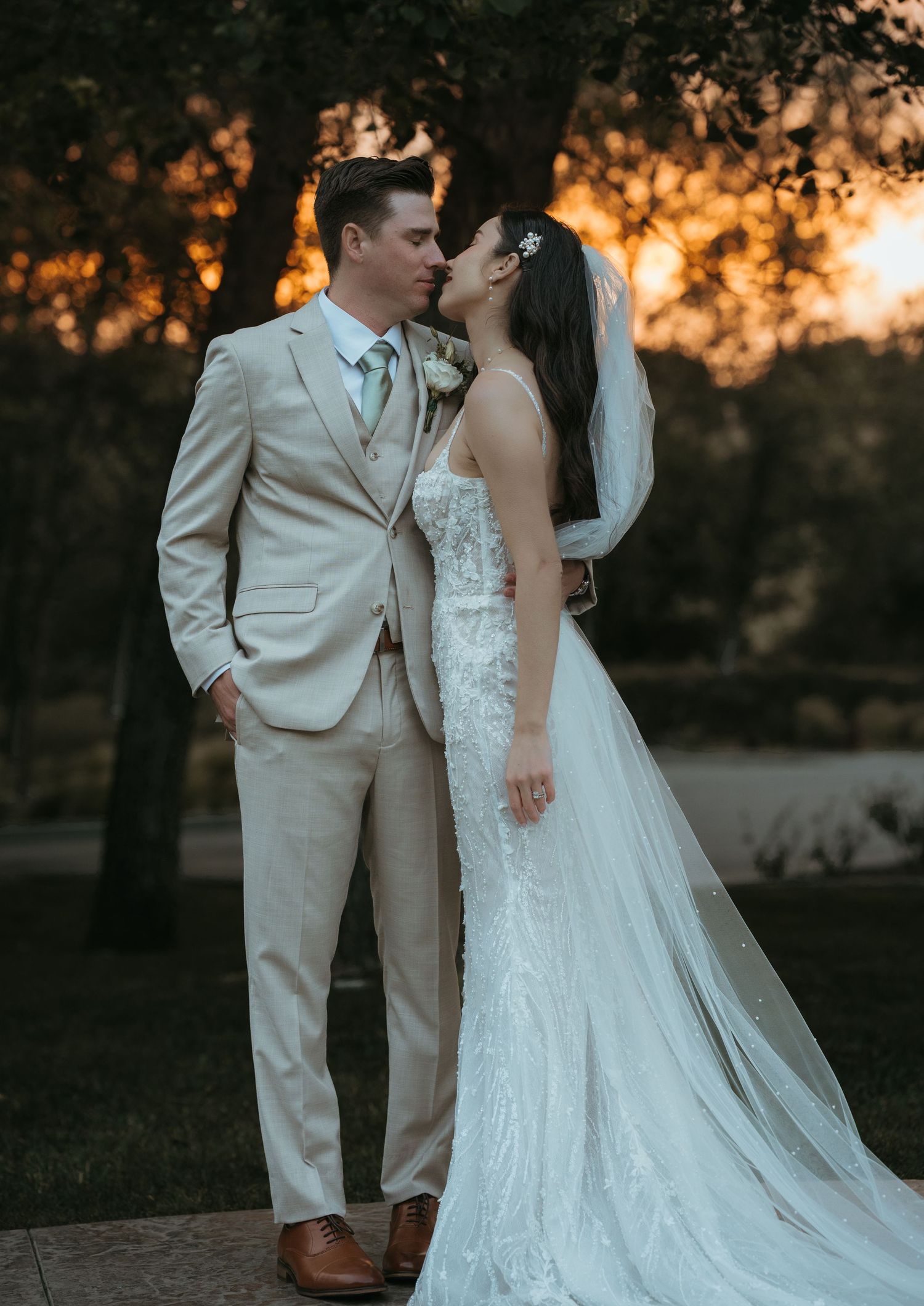 Newlywed couple shares romantic kiss at sunset with glowing orange sky and trees in background.