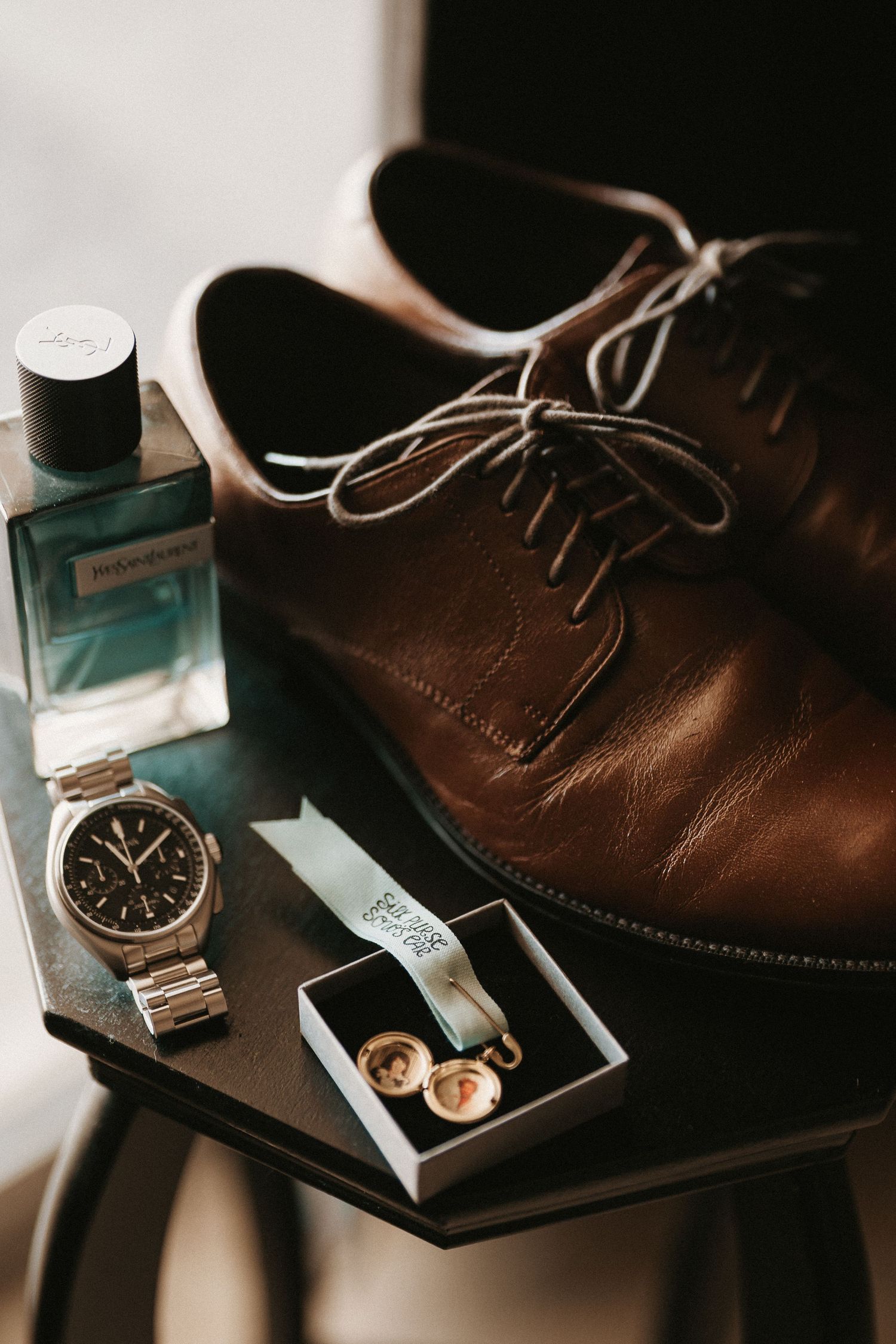 A stylish arrangement of brown leather dress shoes, metal watch, cologne bottle and wedding rings on dark surface.