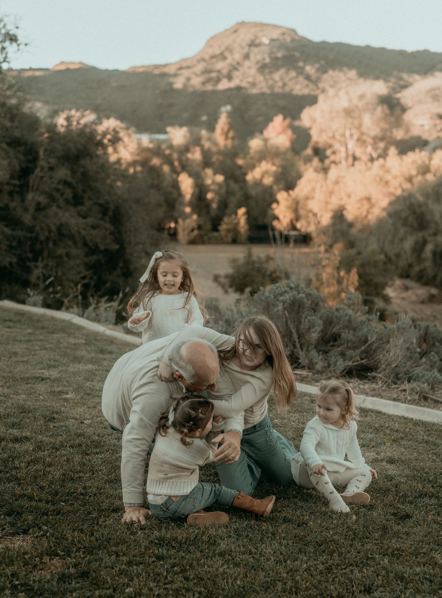 A tender family moment captured on a grassy hillside with mountains visible in the background at sunset.