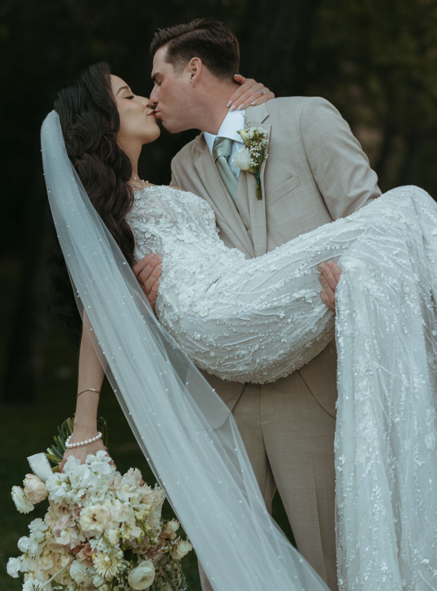 A romantic wedding couple sharing a dip kiss outdoors with flowing white veil and beige suit.