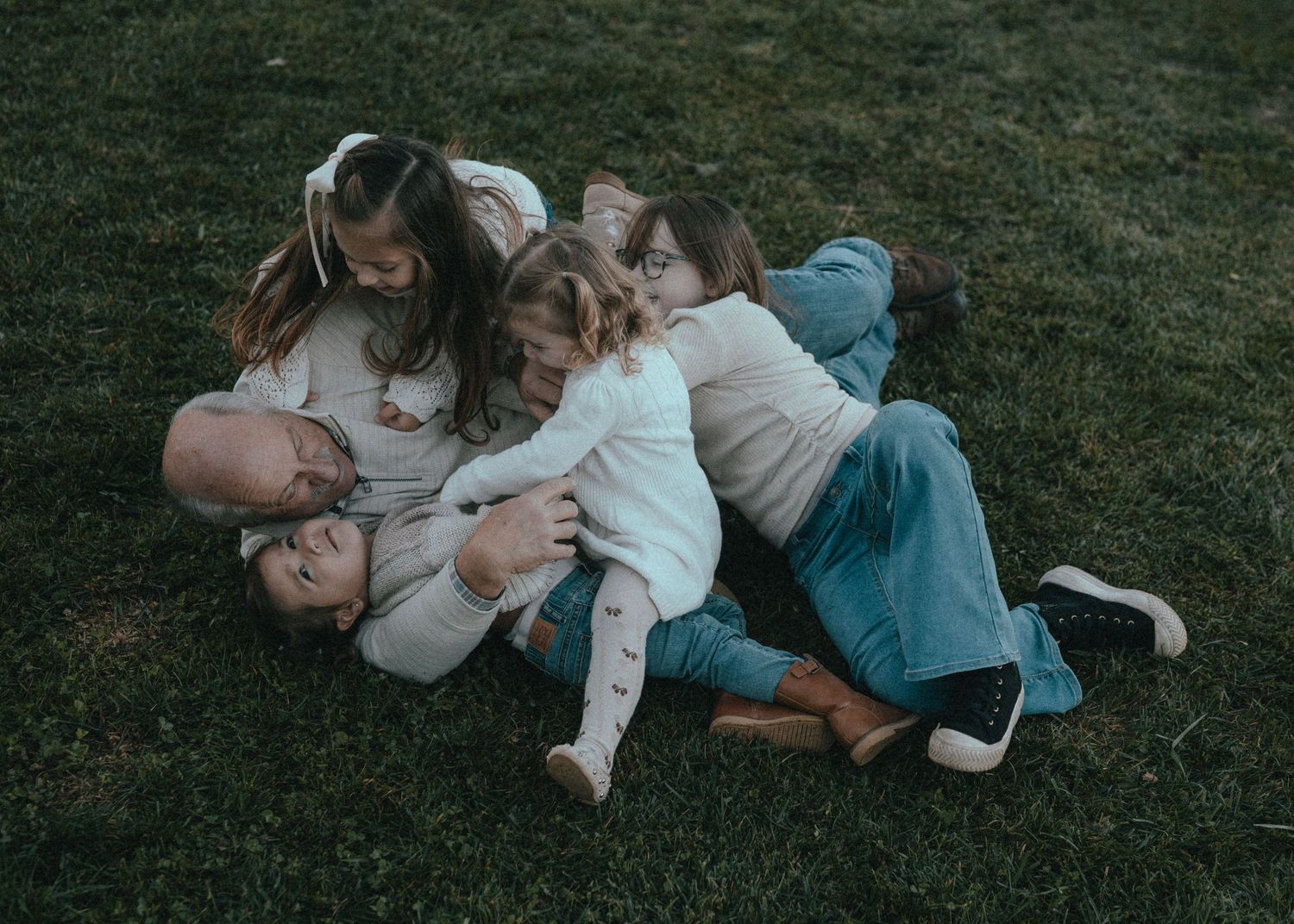 A playful family scene with everyone tackling grandpa down on the grass together in a casual outdoor setting.
