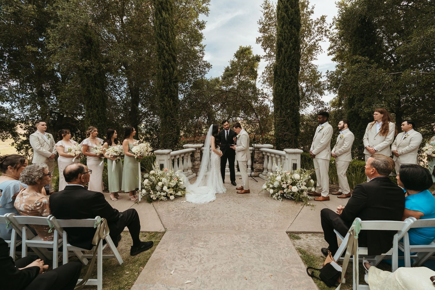 A wedding ceremony takes place outdoors with white chairs arranged in rows and floral decorations lining the aisle.
