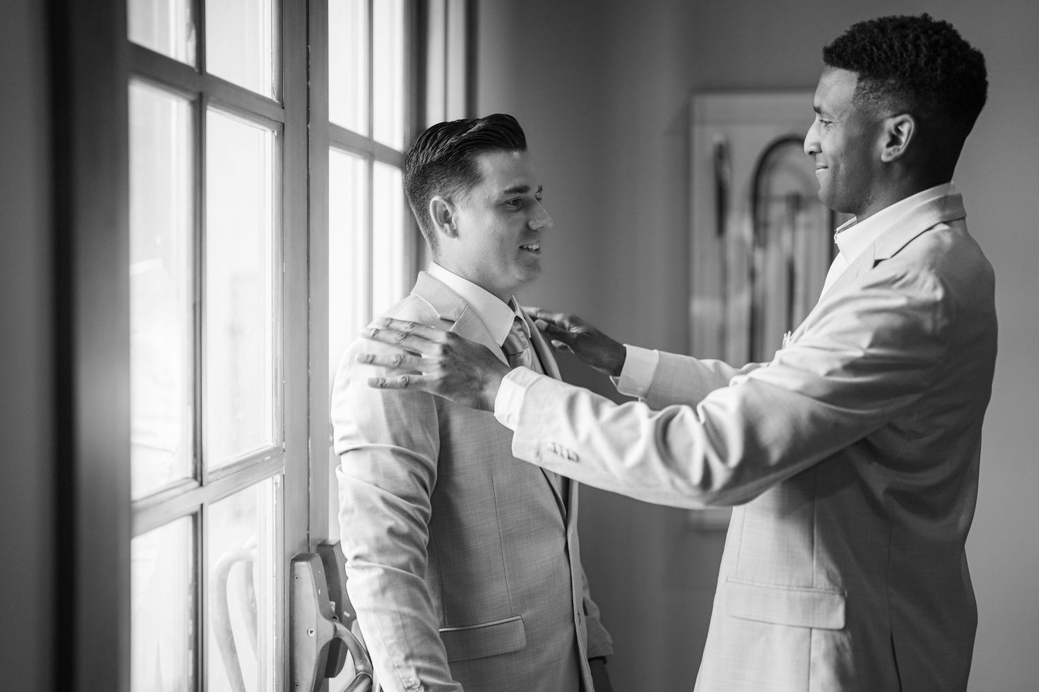 Black and white photo of two men in white suits, one adjusting the other's bow tie near a window.