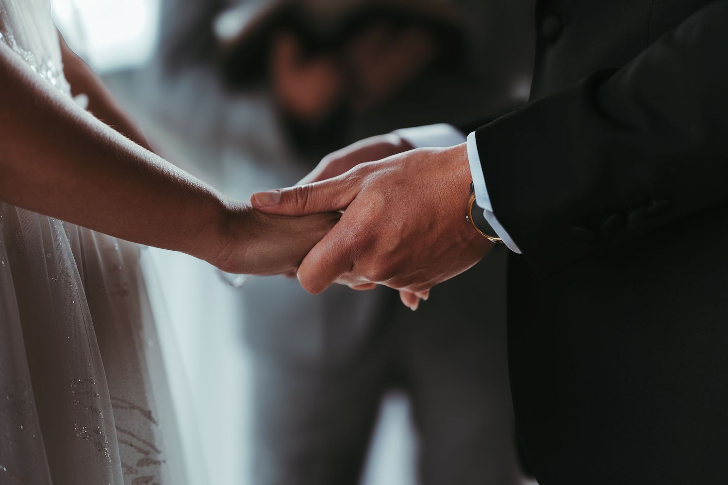 Close-up view of hands clasping together during a wedding ceremony, with white dress and dark suit visible.