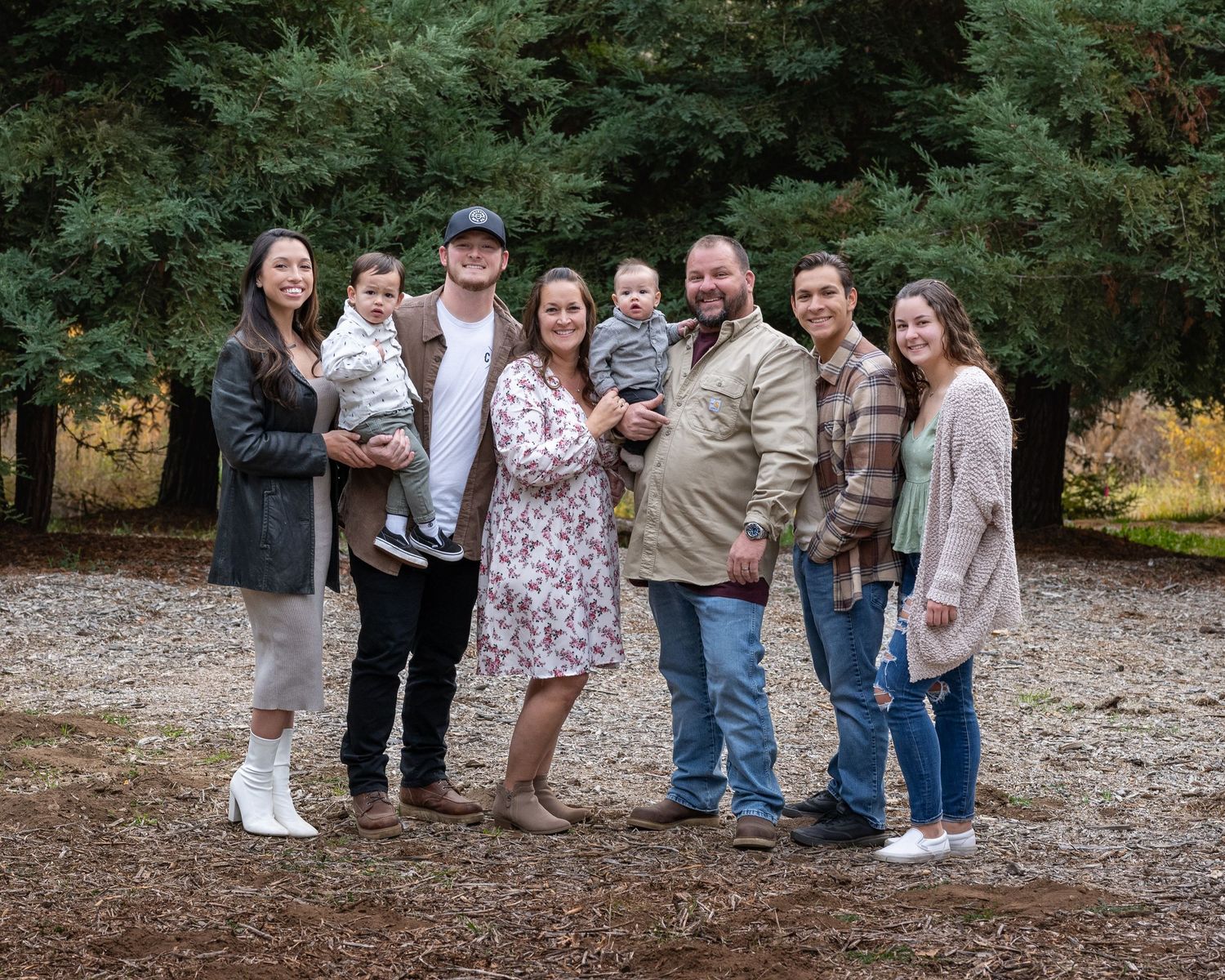 Extended family gathered for outdoor photo session among evergreen trees.