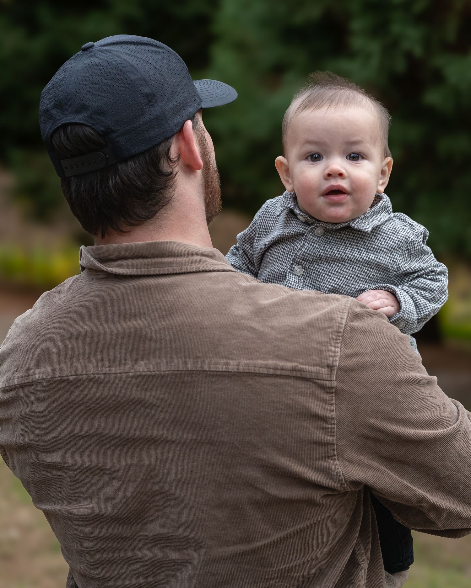 Adult in brown jacket and baseball cap holding a baby outdoors.