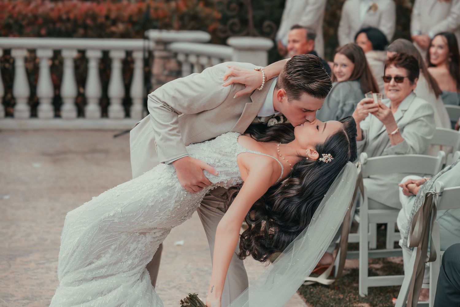 Couple shares a romantic kiss during their wedding ceremony while guests look on at an outdoor venue.
