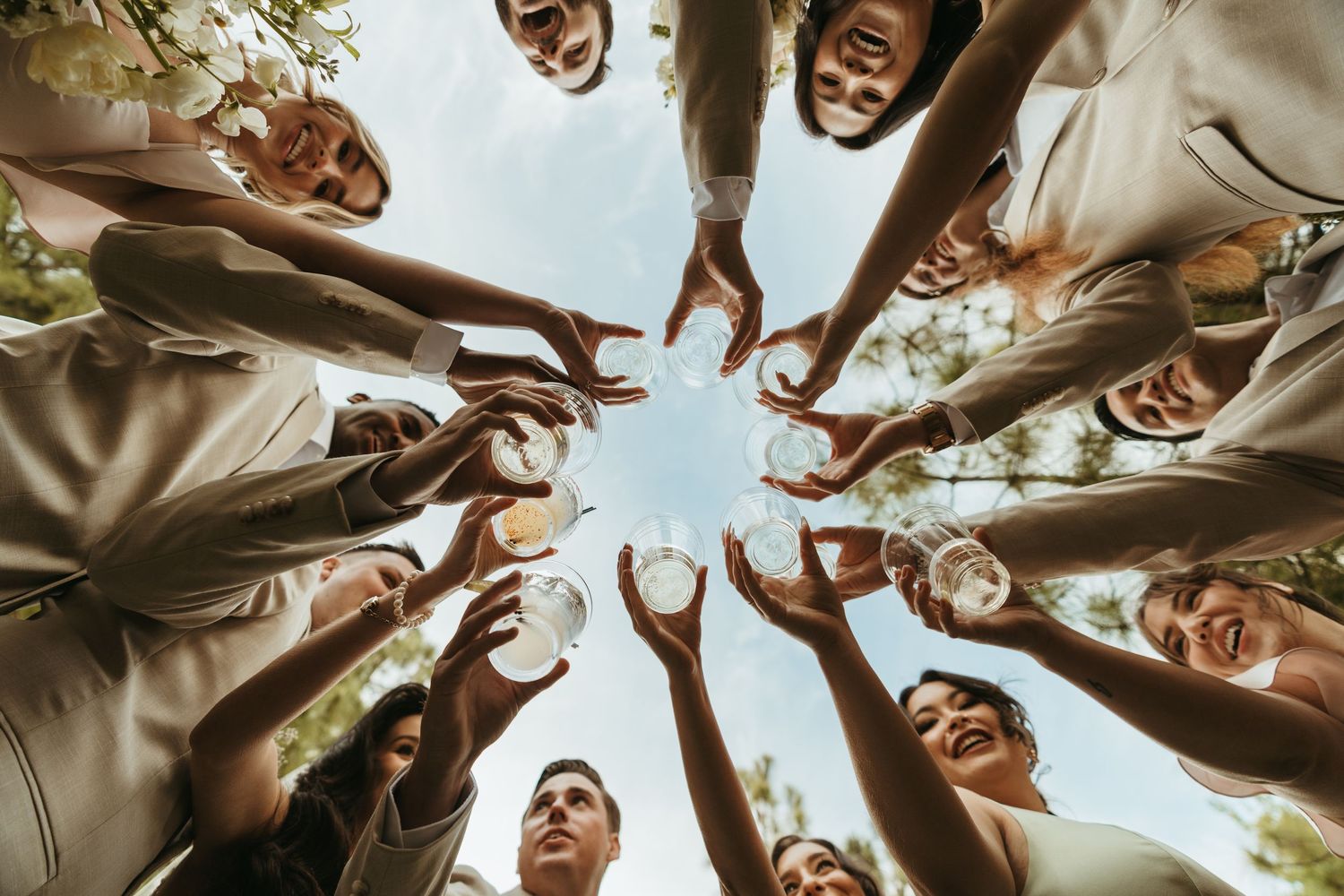 A group of people raise their glasses in a celebratory toast from a low-angle perspective against a bright sky.