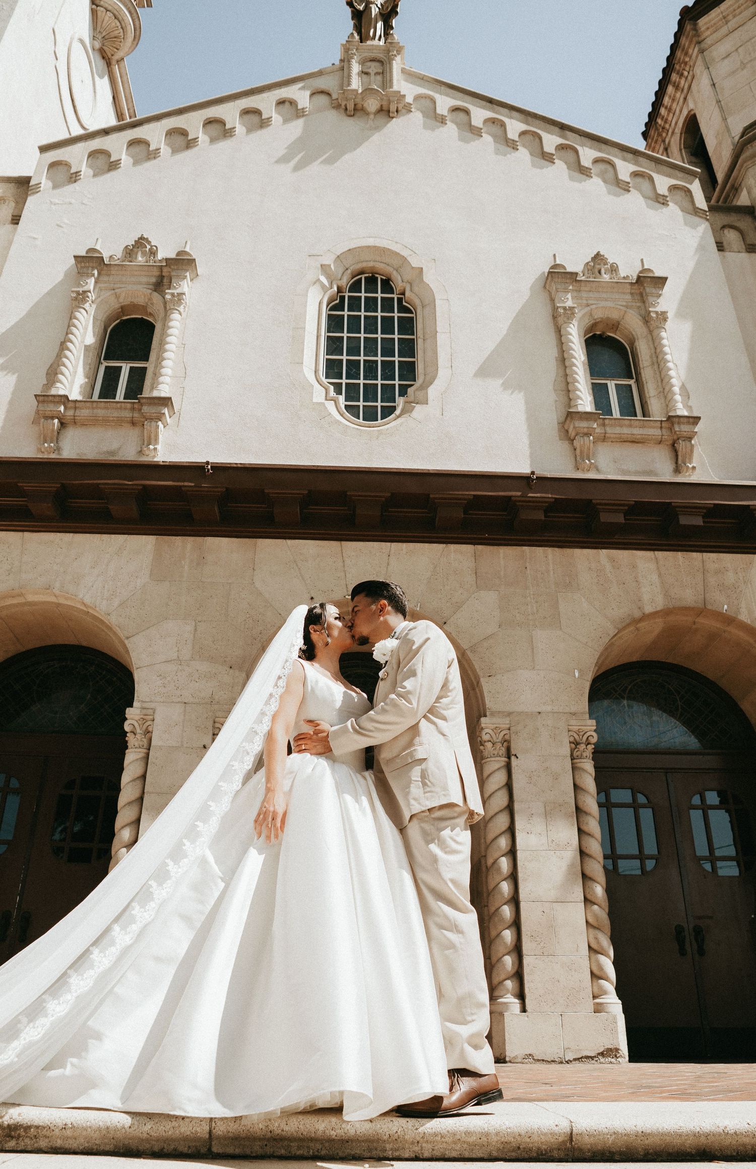A couple shares a romantic moment on the steps of an elegant white church with ornate architectural details and arched windows.