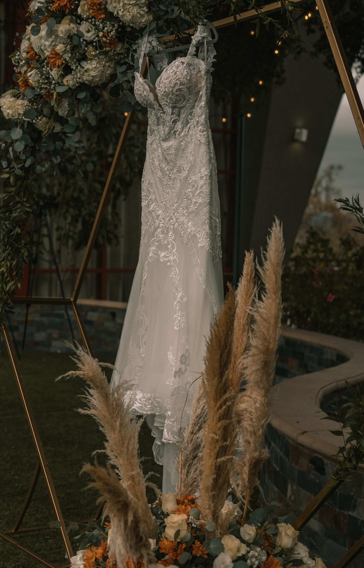 Elegant white wedding dress with lace details displayed against dried pampas grass and floral decorations.