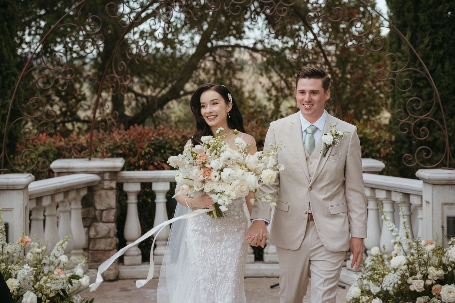 A couple in wedding attire stands on a stone balcony with floral arrangements in a garden setting.
