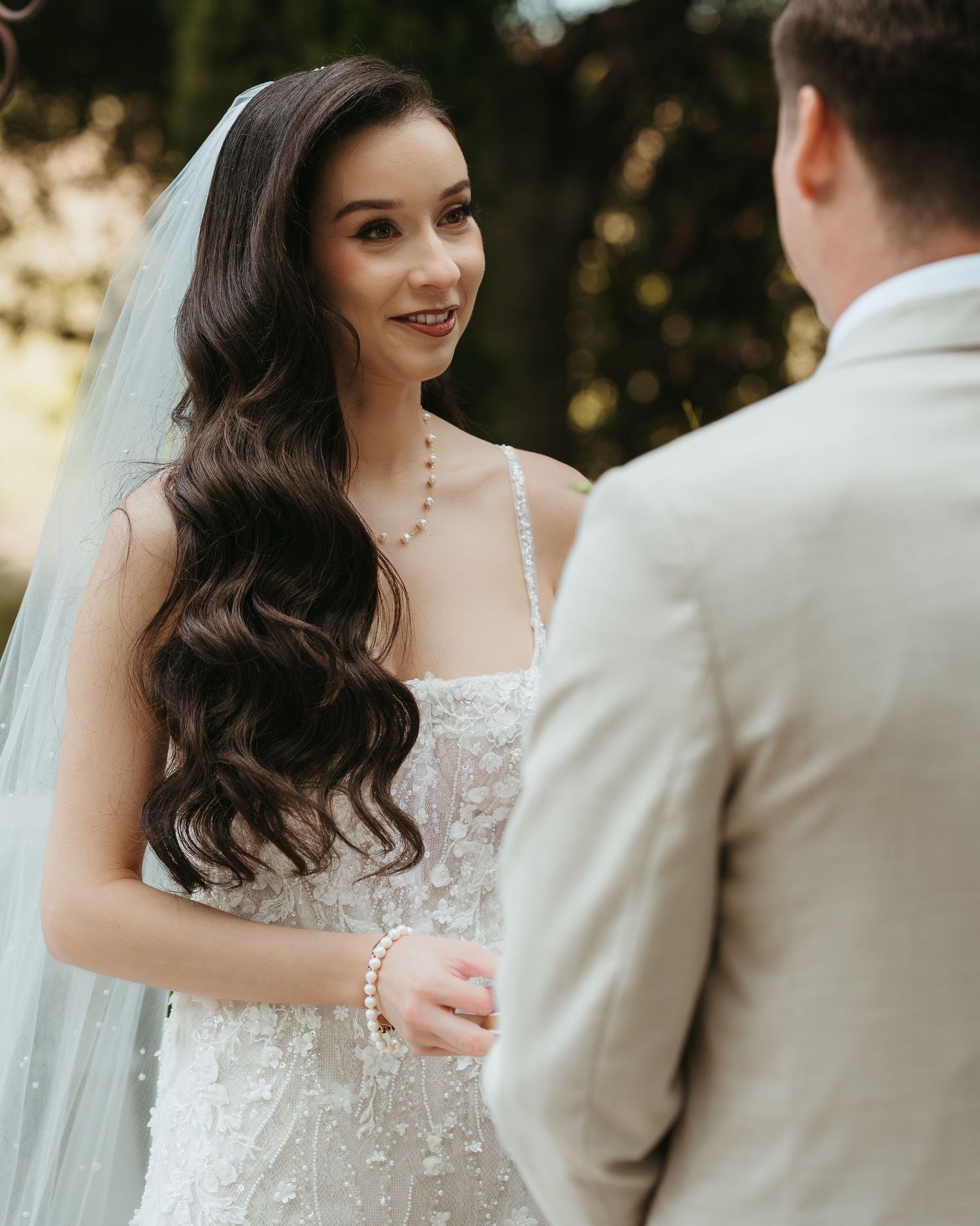 A bride in a lace wedding dress with long wavy hair smiles during an outdoor wedding ceremony.