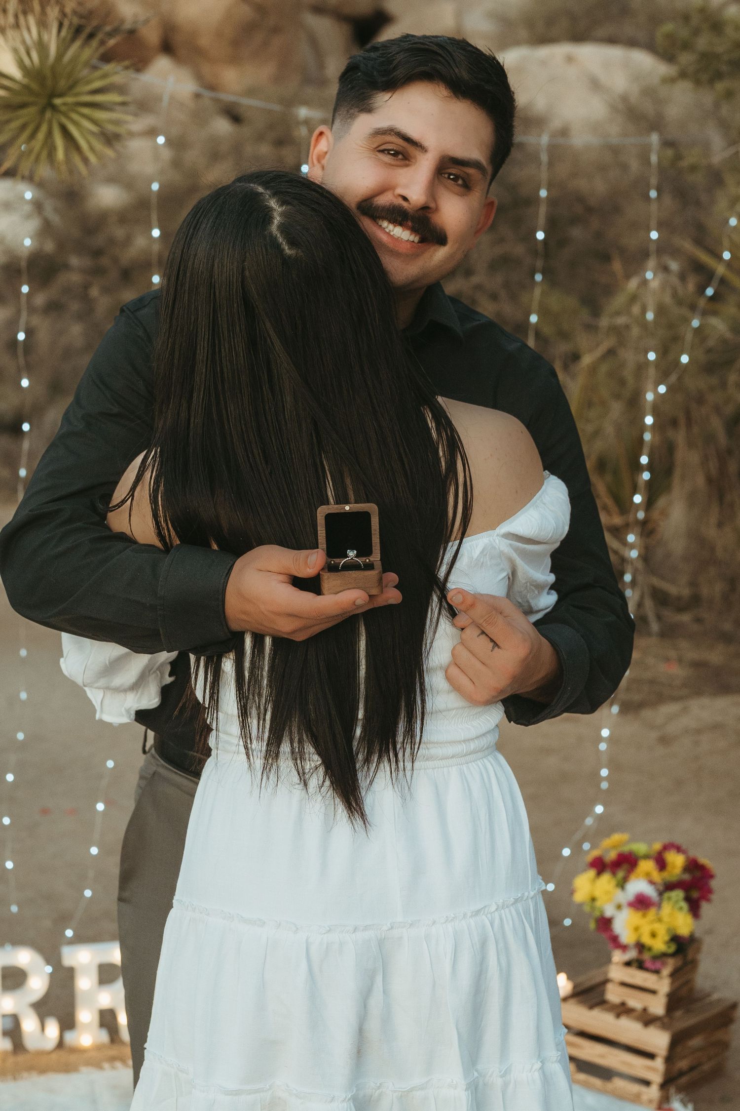 A romantic proposal moment decorated with string lights and wooden crates in a desert setting.