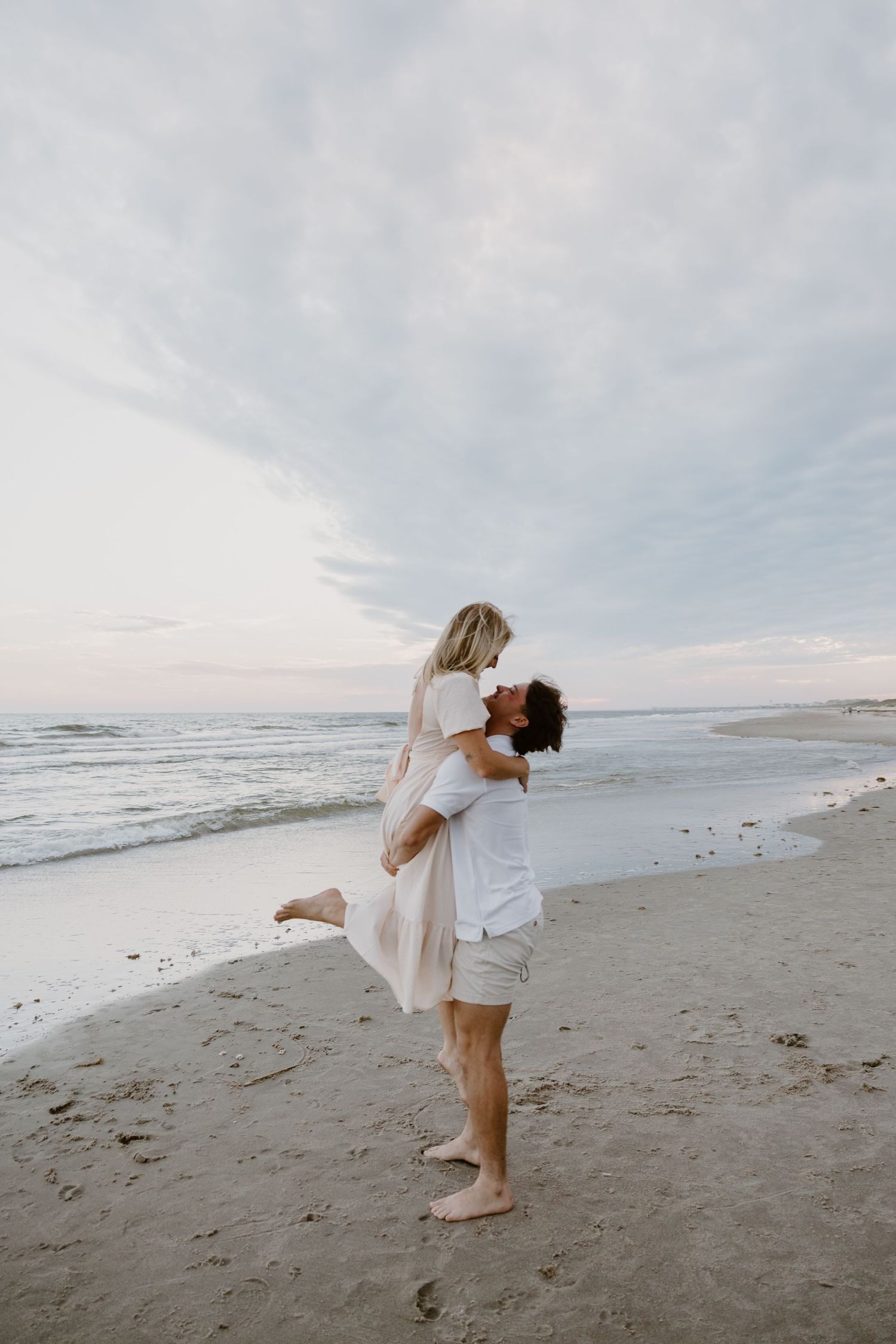 A couple in white clothing dances playfully on a beach at sunset.