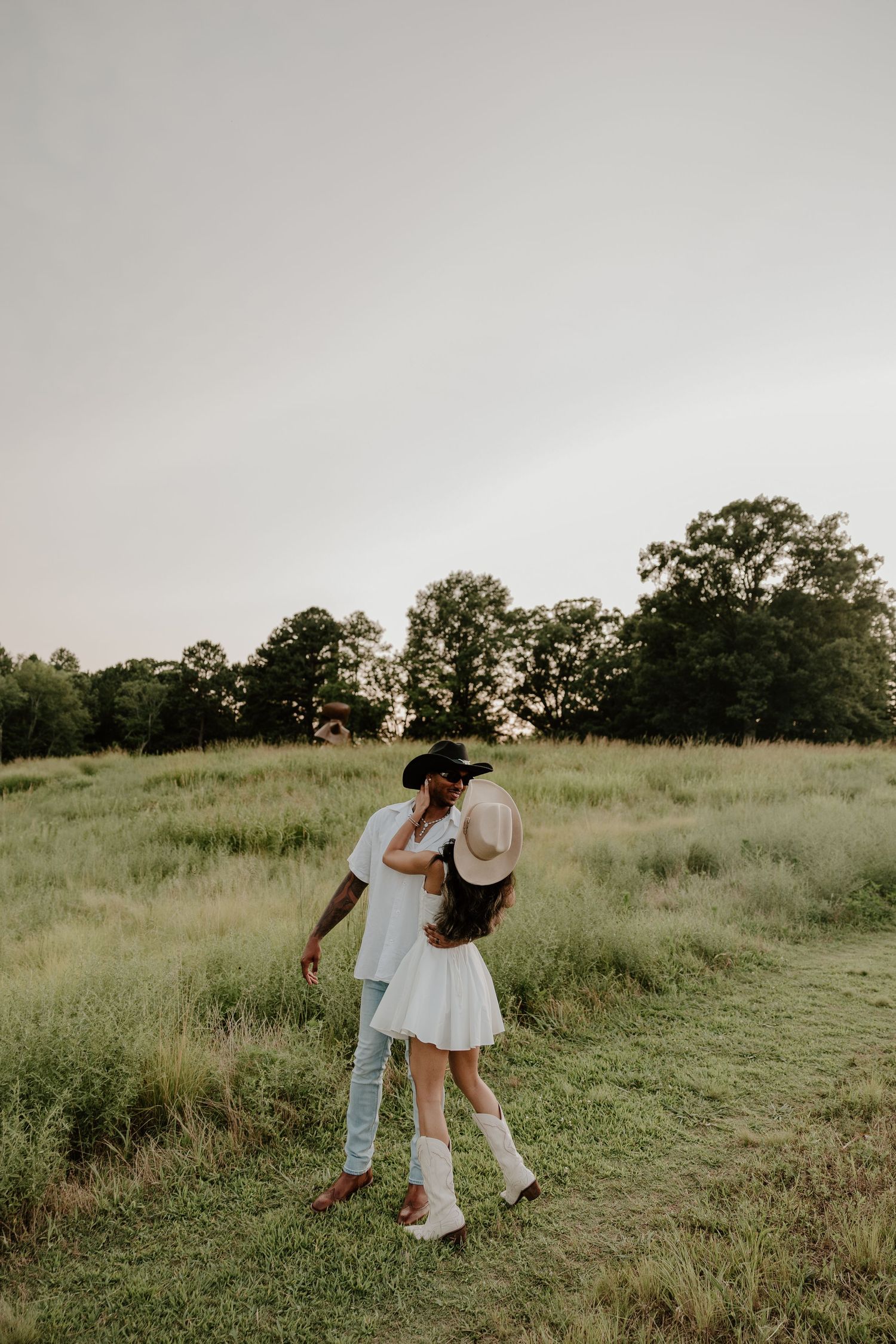 Romantic couple in white clothing playfully embracing on a grassy hillside at dusk.