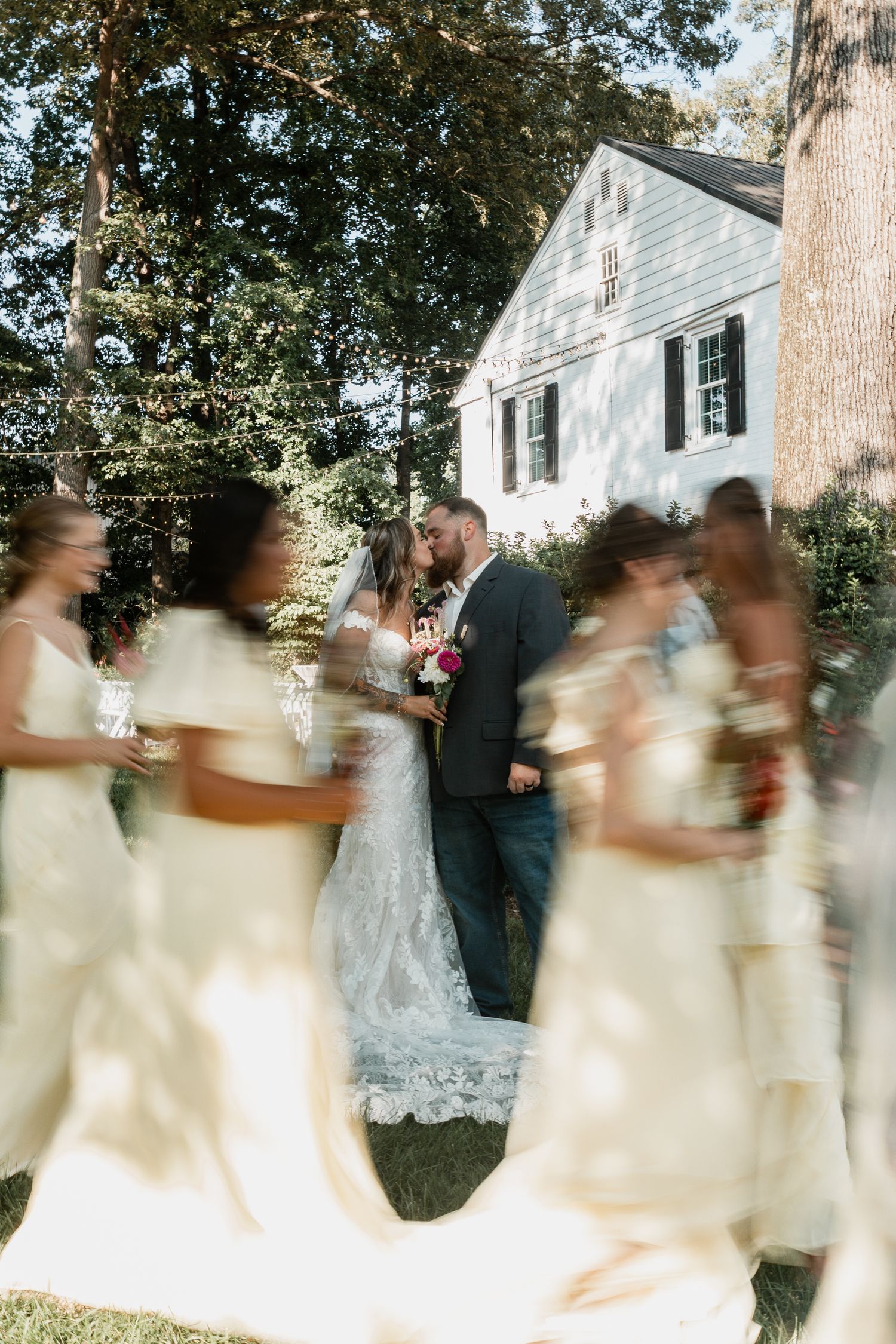 Motion blur of bridesmaids in cream dresses walking past white house during wedding.