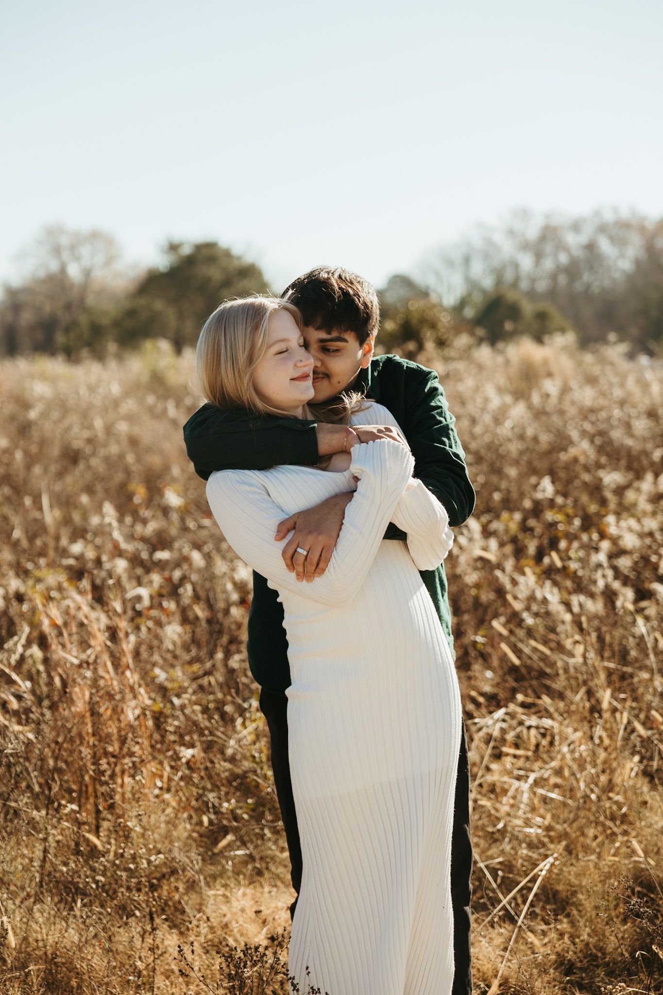 A couple embraces in a golden field during autumn wearing white and dark sweaters.