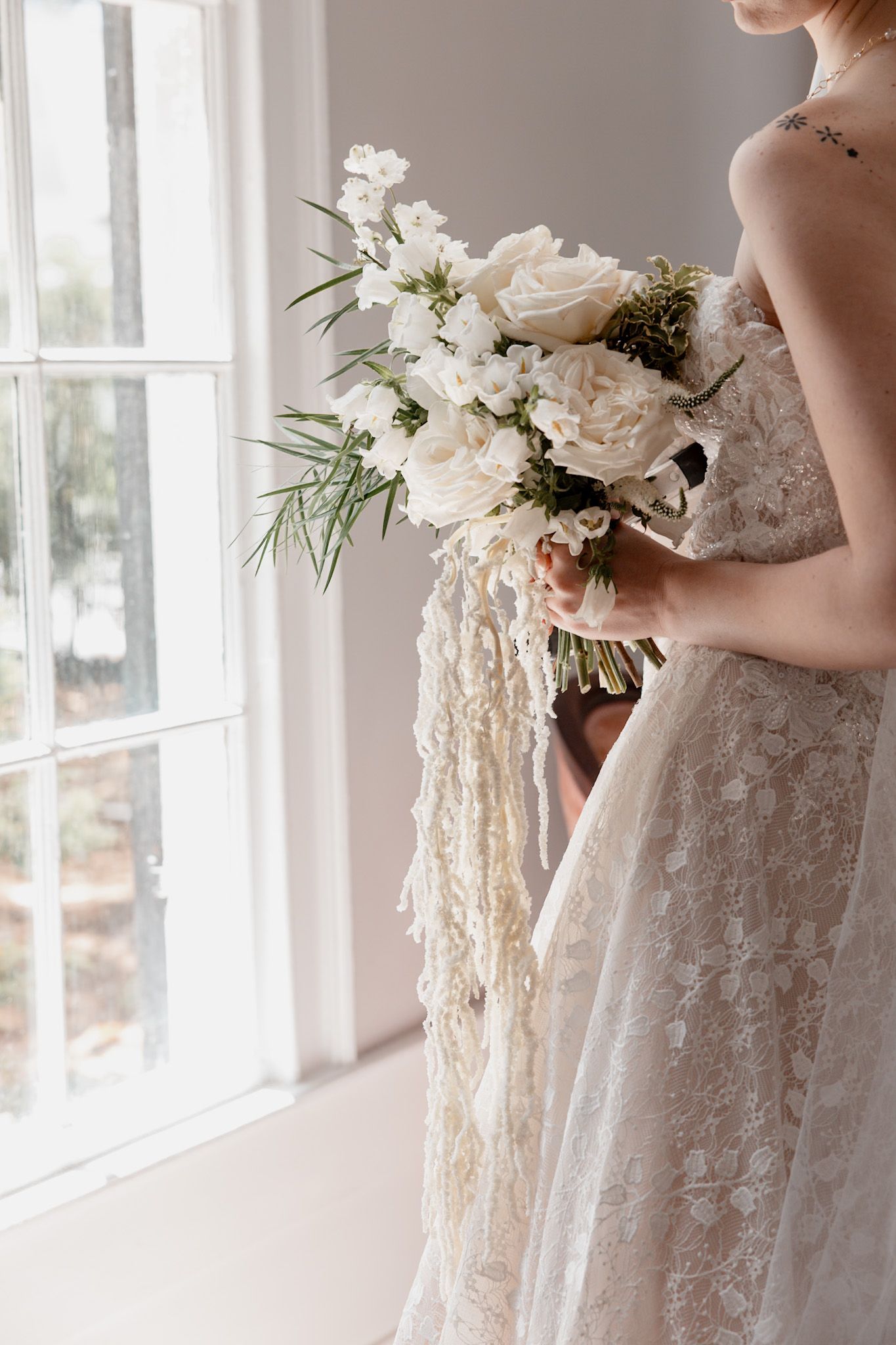 Elegant white floral bouquet with cascading lace ribbons held against a lace wedding dress by a window.