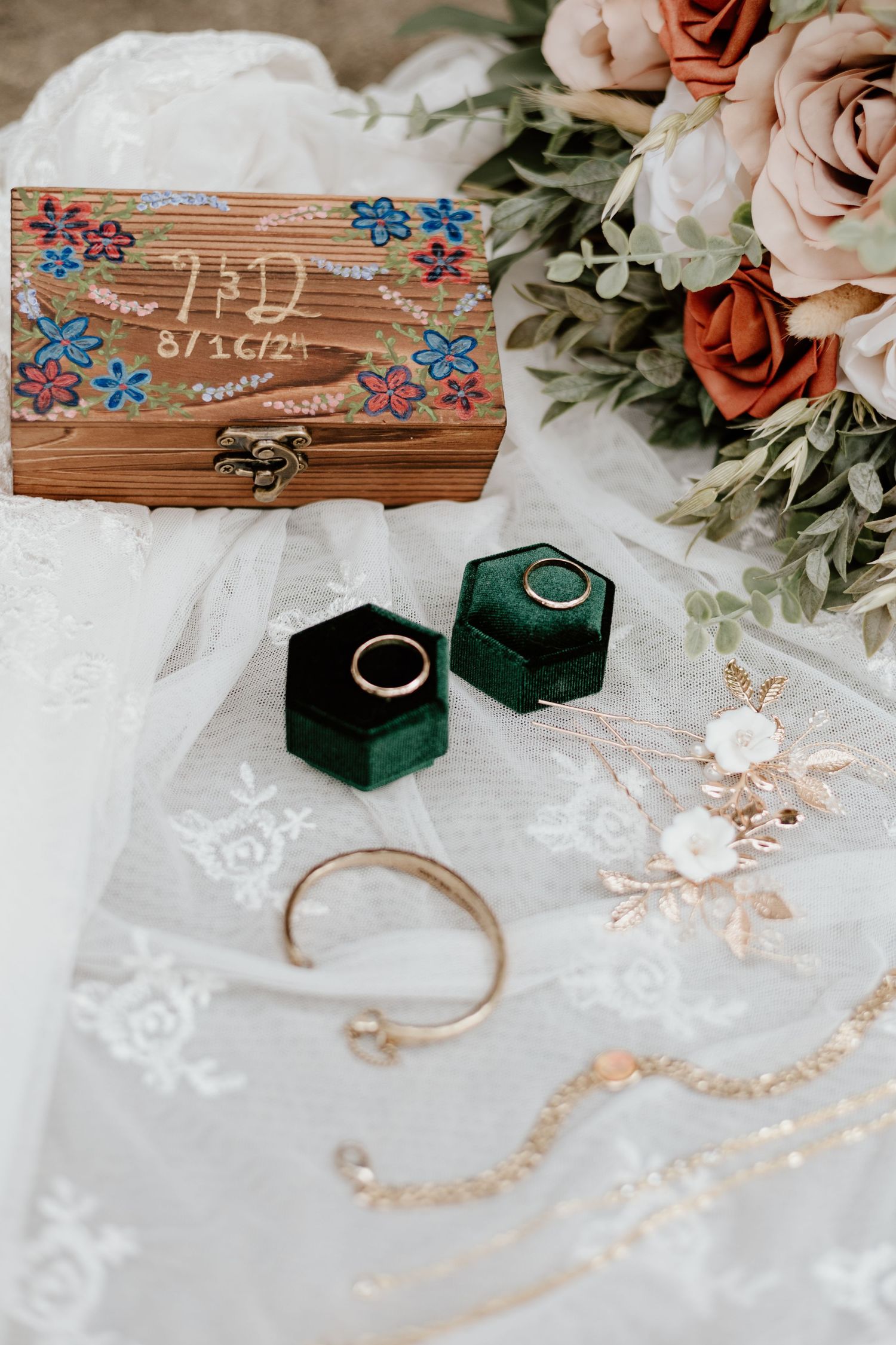 Wedding rings in green velvet boxes with wooden floral box and roses on lace fabric.