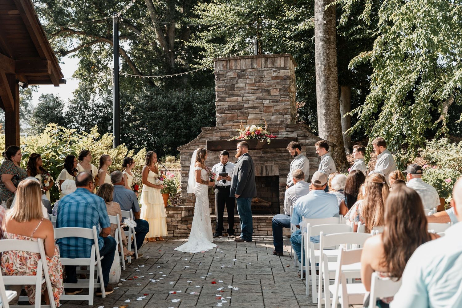 Outdoor wedding ceremony with stone fireplace backdrop and white chairs on stone patio.