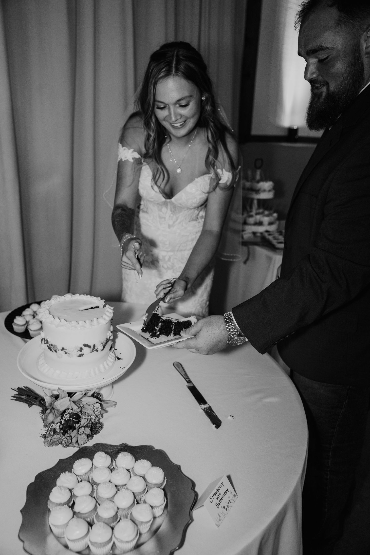 Black and white image of a wedding cake cutting ceremony with cupcakes displayed on a white tablecloth.