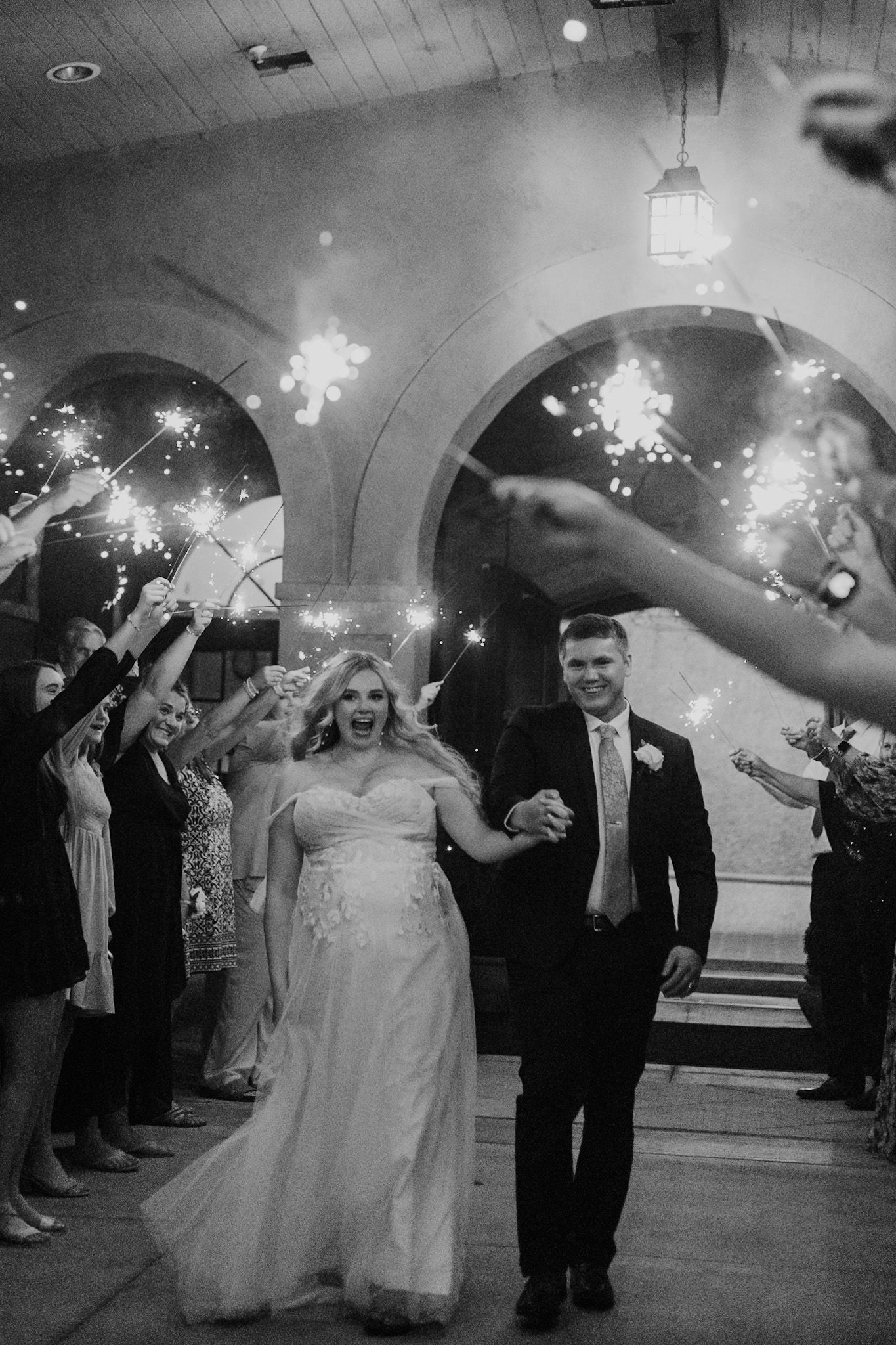Wedding couple exits reception under a glowing sparkler arch held by guests in a dimly lit venue.