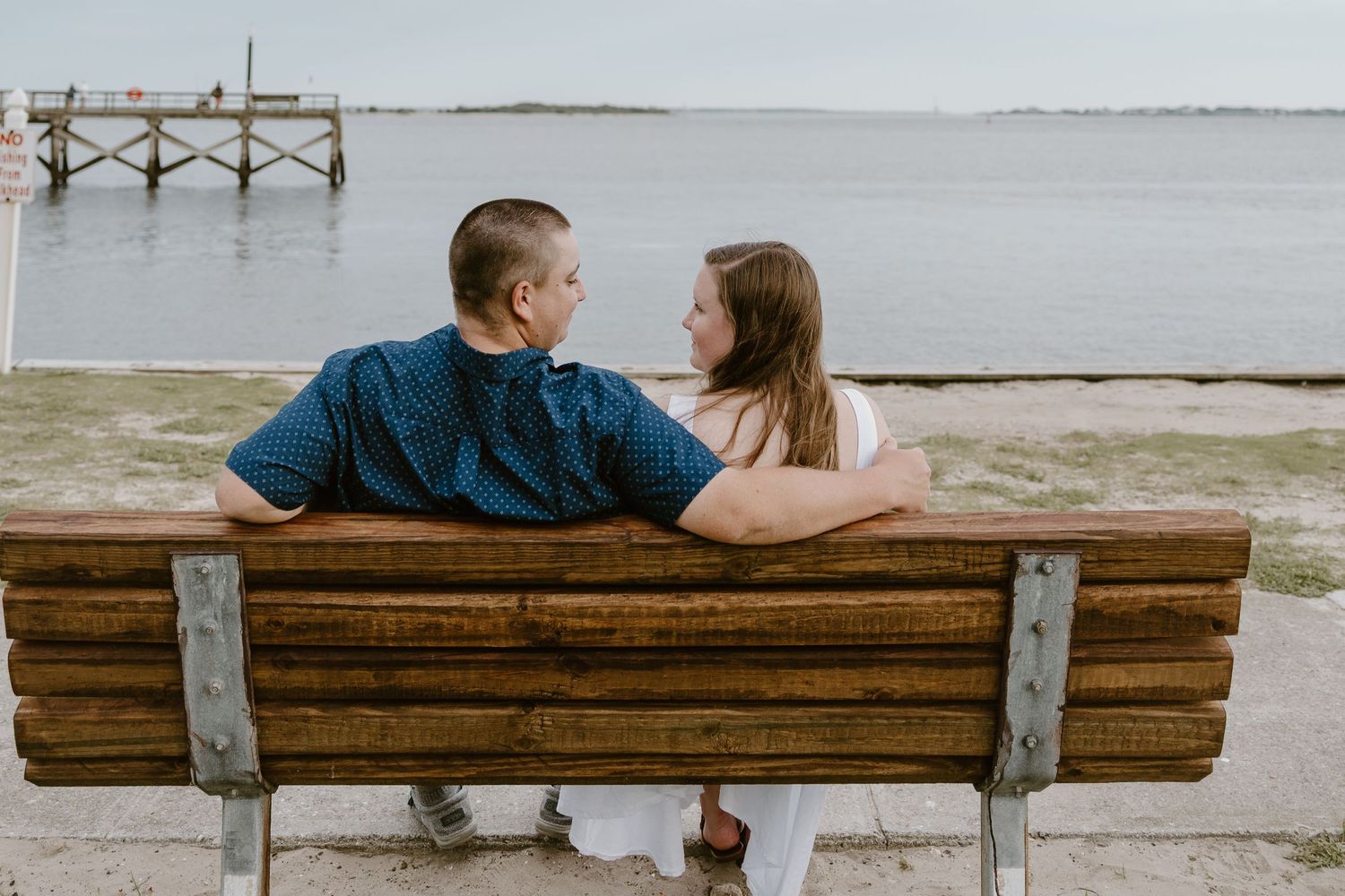 Couple sharing intimate moment while sitting on wooden bench overlooking waterfront dock.