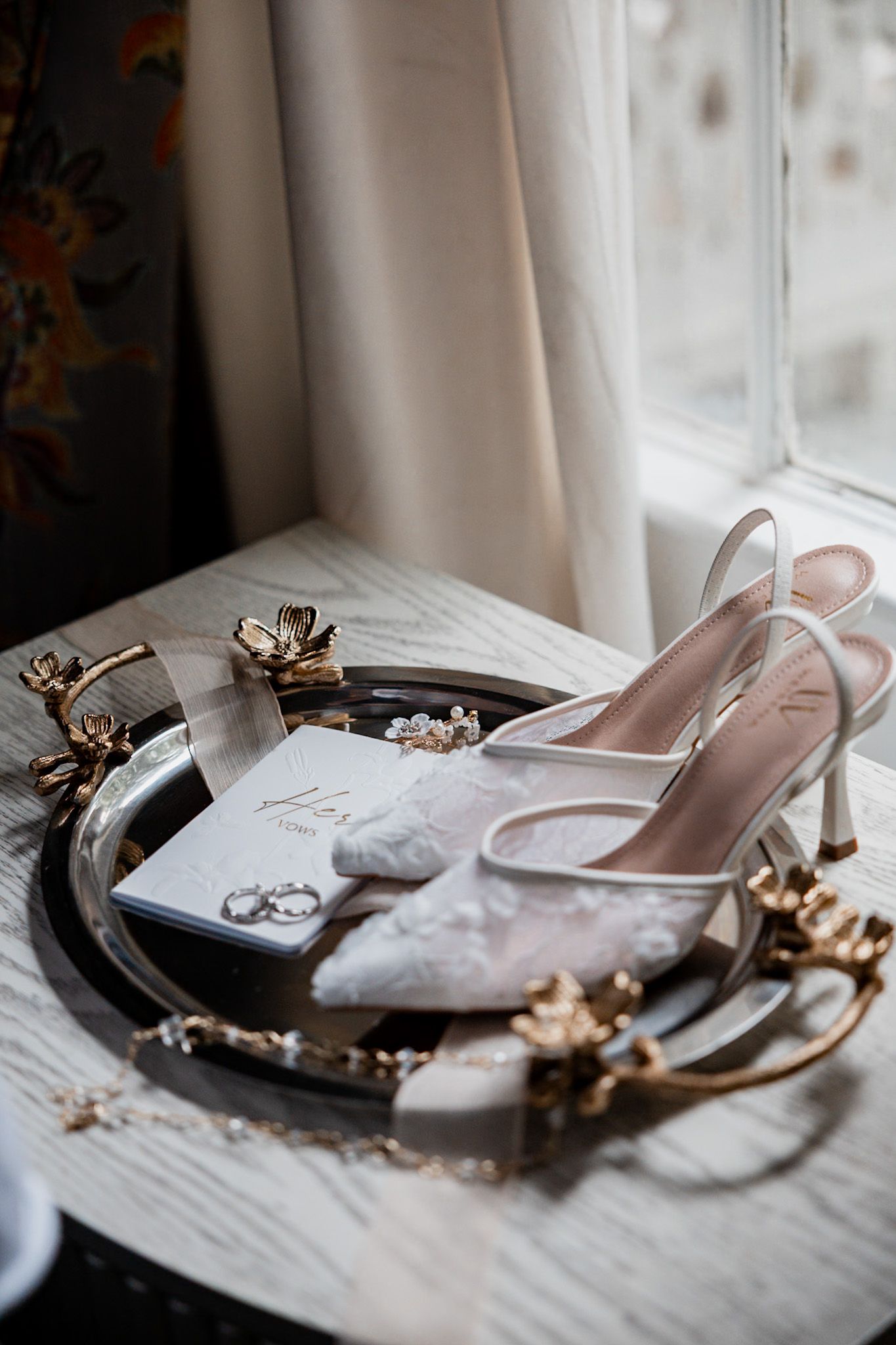 Wedding accessories including silver shoes and jewelry arranged on a windowsill with natural light.