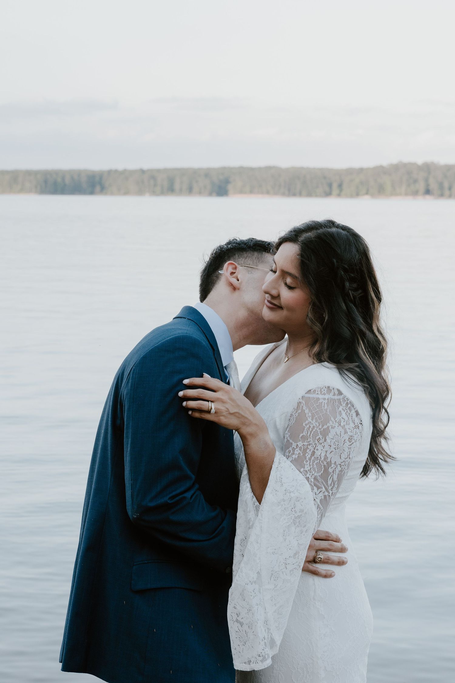 Intimate waterfront moment between couple in formal attire with lake and forest in background.