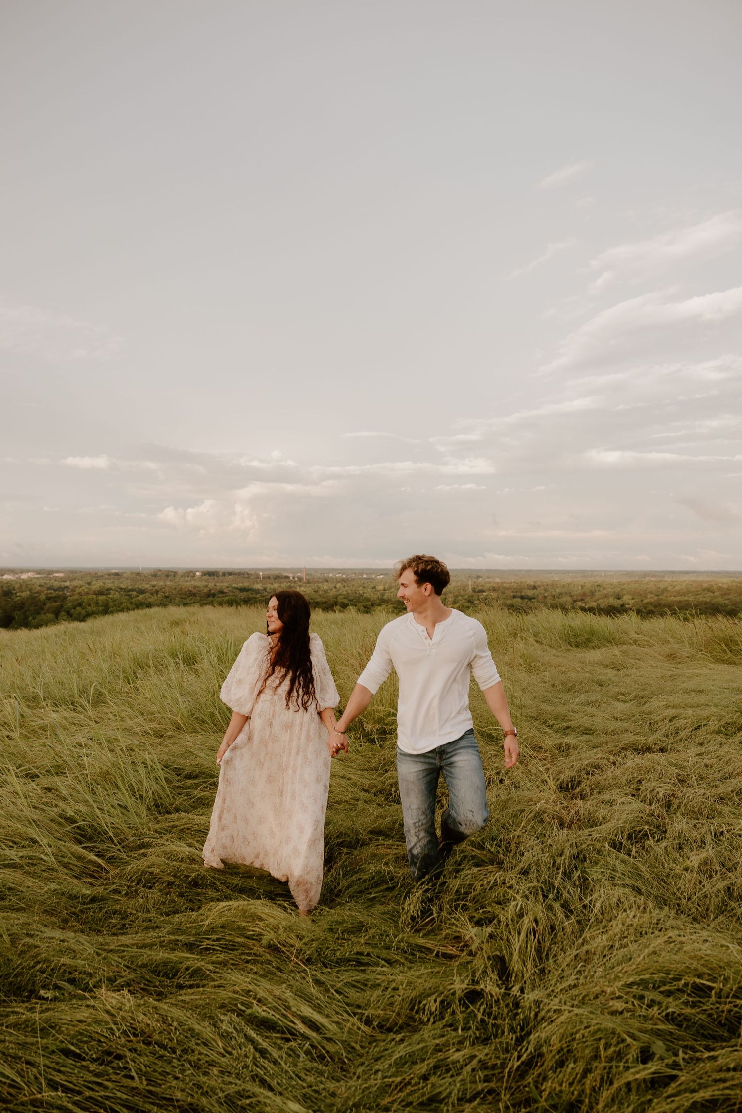 Couple walking hand in hand through tall grass in a golden field at sunset.